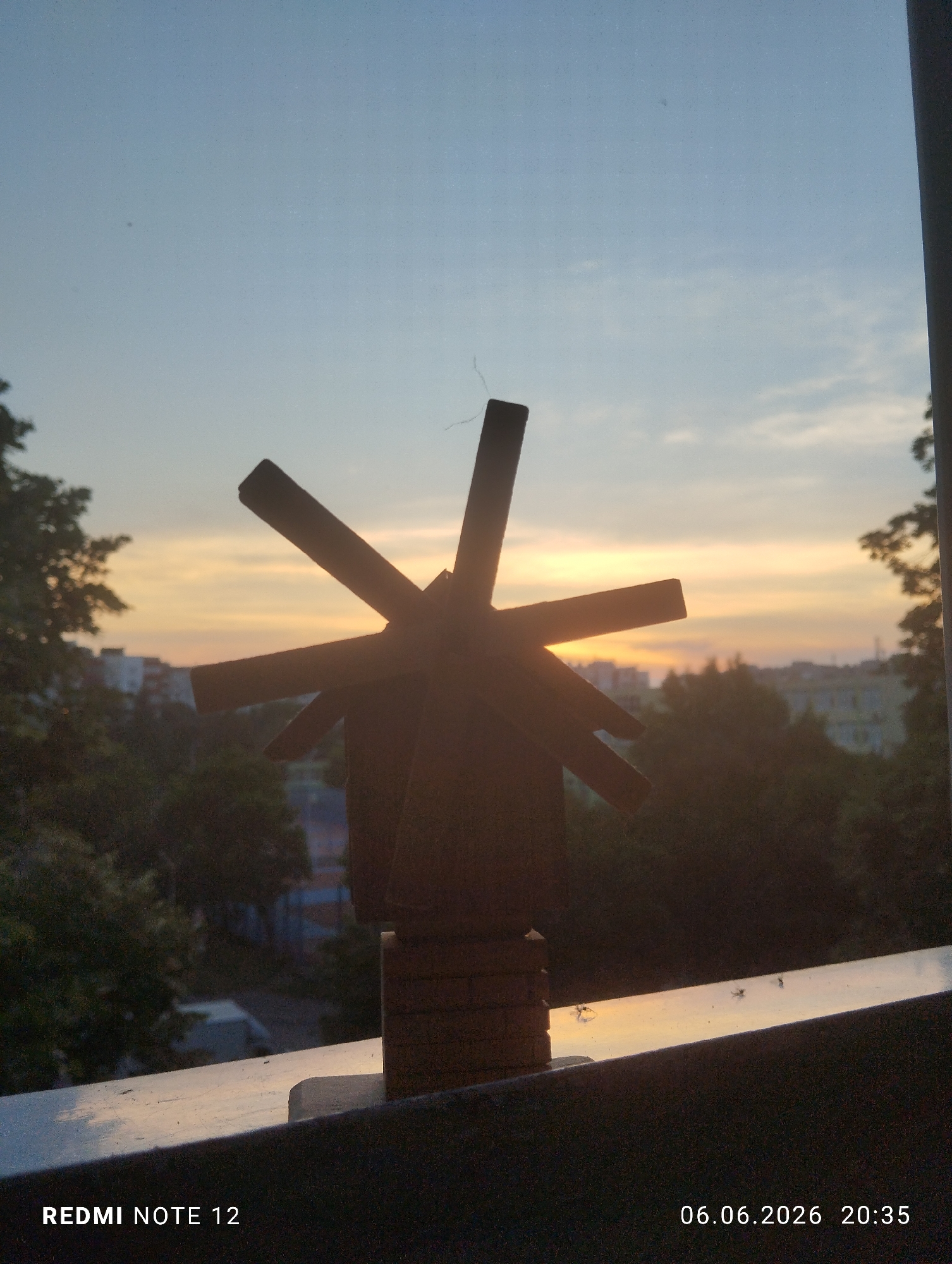A photograph taken on June 6, 2026, at 20:35, depicts a miniature wooden windmill model silhouetted against a sunset in Burgas, Bulgaria. The windmill, featuring four blades and a visible brick-patterned base, stands on a light-colored outdoor surface, likely a balcony railing, which exhibits minor debris. The strong backlighting from the setting sun, positioned directly behind the windmill, creates a dramatic silhouette effect. The sky transitions from a bright, golden-orange hue at the horizon to a lighter blue overhead, with minimal cloud cover. In the midground, dense green tree foliage partially obscures several multi-story residential buildings, indicating an urban landscape. The image includes a "REDMI NOTE 12" watermark in the bottom-left corner.