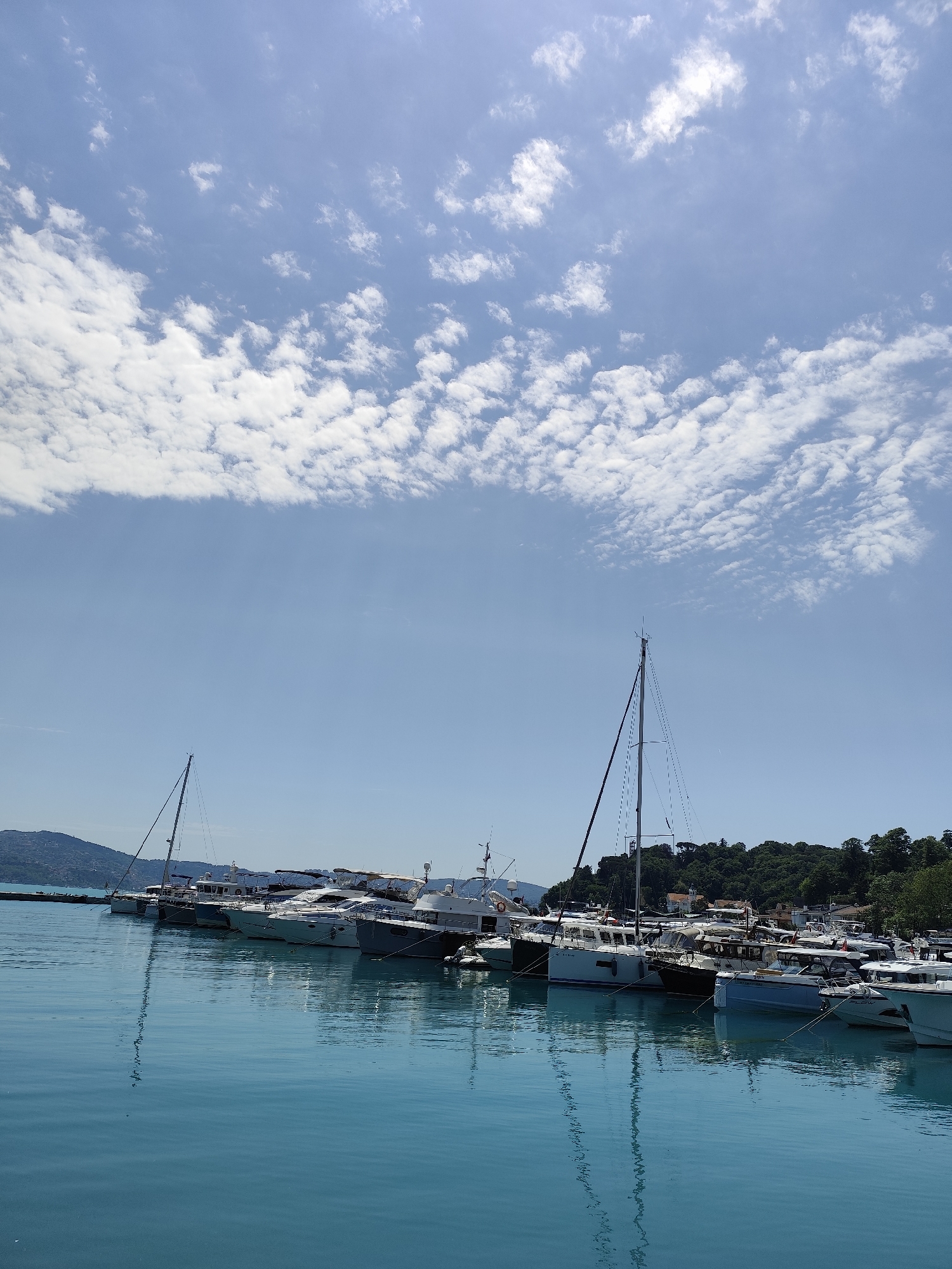 The image displays a tranquil daytime scene at a marina in Sarıyer, Turkey. Numerous boats, including large yachts and several sailboats, are docked along a pier. Their hulls range in color from white to dark blue, with various coverings and masts visible. The prominent mast of a sailboat stands tall towards the center-right of the frame.
The water in the foreground is a striking turquoise-blue, appearing calm and highly reflective, mirroring the boats and the sky above with gentle ripples. In the background, a lush, tree-covered hill extends on the right, providing a natural backdrop to the marina. To the left, a distant mountainous coastline is visible under the bright sky.
The sky occupies a significant portion of the image, showing a clear, light blue expanse adorned with scattered white cumulus clouds. The sunlight is bright, suggesting a sunny and pleasant day. No people are visible in the scene, and there is no discernible text on any of the boats or structures. The overall atmosphere conveys a sense of calm and leisure in a scenic coastal setting.