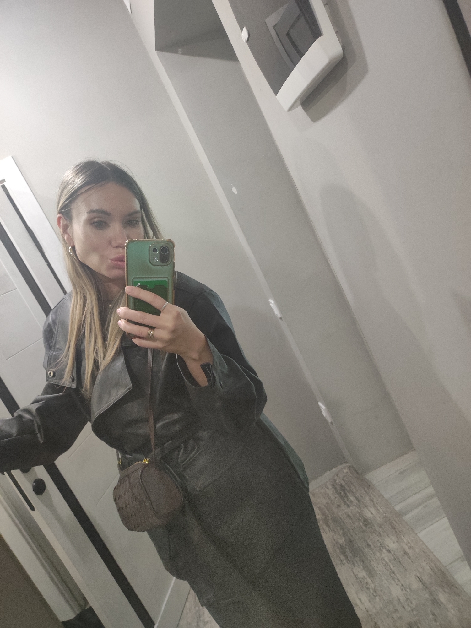 A woman stands in an interior space, likely a restroom or hallway, taking a selfie in a mirror. She is wearing a dark leather-like outfit, possibly a trench coat and skirt or dress, and a small brown cross-body bag. Her hair is blonde with darker roots and she is wearing hoop earrings and rings. The background consists of a light-colored wall, a door with black trim, and what appears to be a white fixture mounted on the wall. The floor is visible and appears to be made of light-colored material. There is no indication that the image was taken in Votkinsk, Russia.