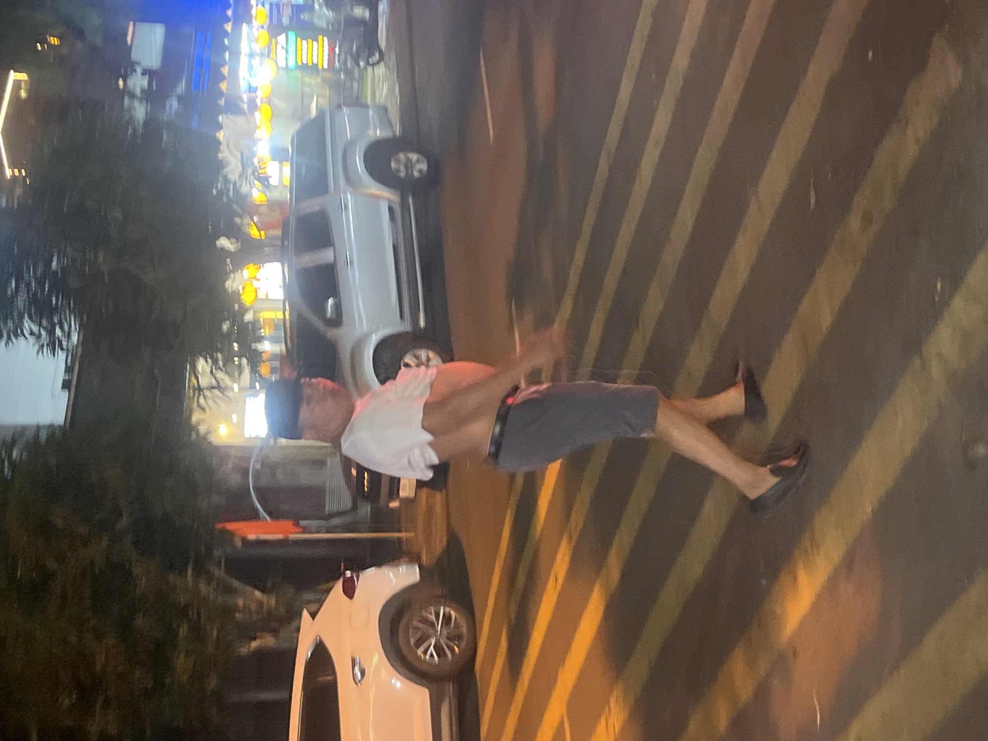 A man wearing a white t-shirt, dark shorts, and black loafers walks across a crosswalk at night. The man is wearing a dark cap. The crosswalk has yellow and dark stripes. In the background, there is a white SUV on the left and a silver pickup truck on the right. Buildings with illuminated signs are visible in the distance, suggesting an urban, possibly commercial area. The lighting indicates it is nighttime.