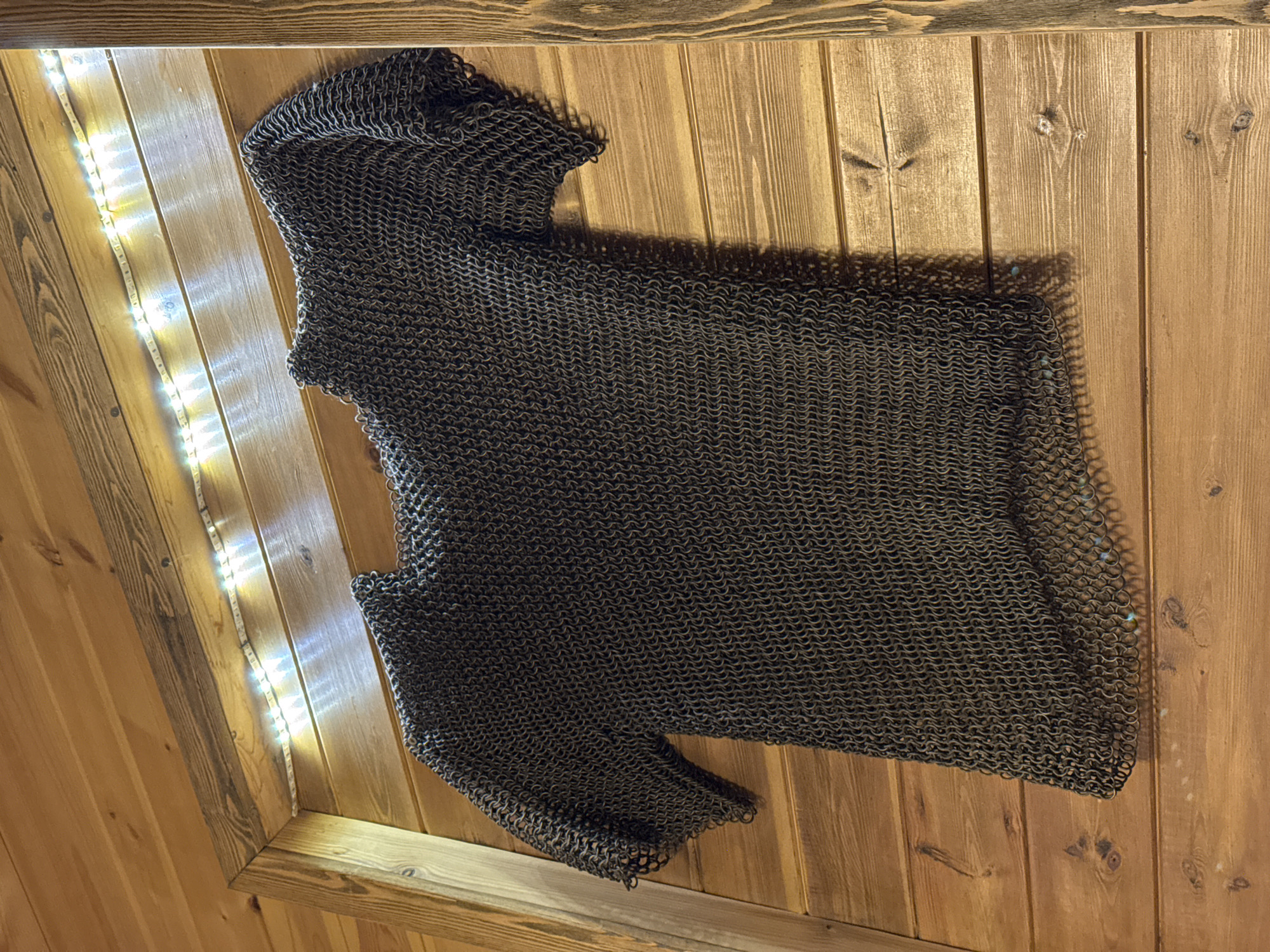 The image depicts a chainmail hauberk laid out on a wooden surface. The hauberk appears to be made of metal rings interlinked to form a tunic, with a hood and short sleeves. The wooden surface consists of light-brown, vertically aligned planks. In the upper-left corner, a string of white LED lights is visible. The scene appears to be indoors, with a focus on the hauberk and its texture. There is no indication of the location being Ново-Никольское, Russia, or of any people, actions, or interactions.
