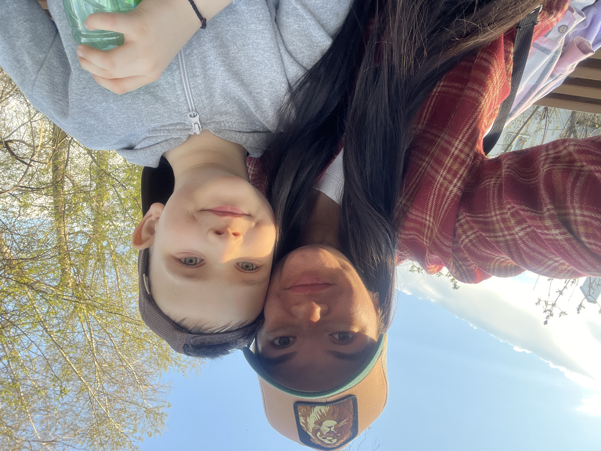 A selfie captures a woman and a child in Kazan, Russia. The woman is wearing a red plaid shirt and a tan baseball cap with a lion patch. The child, to her left, is wearing a gray hooded sweatshirt and a dark-colored cap. Both are looking at the camera with slight smiles. The background features blue sky and tree branches with new spring leaves.