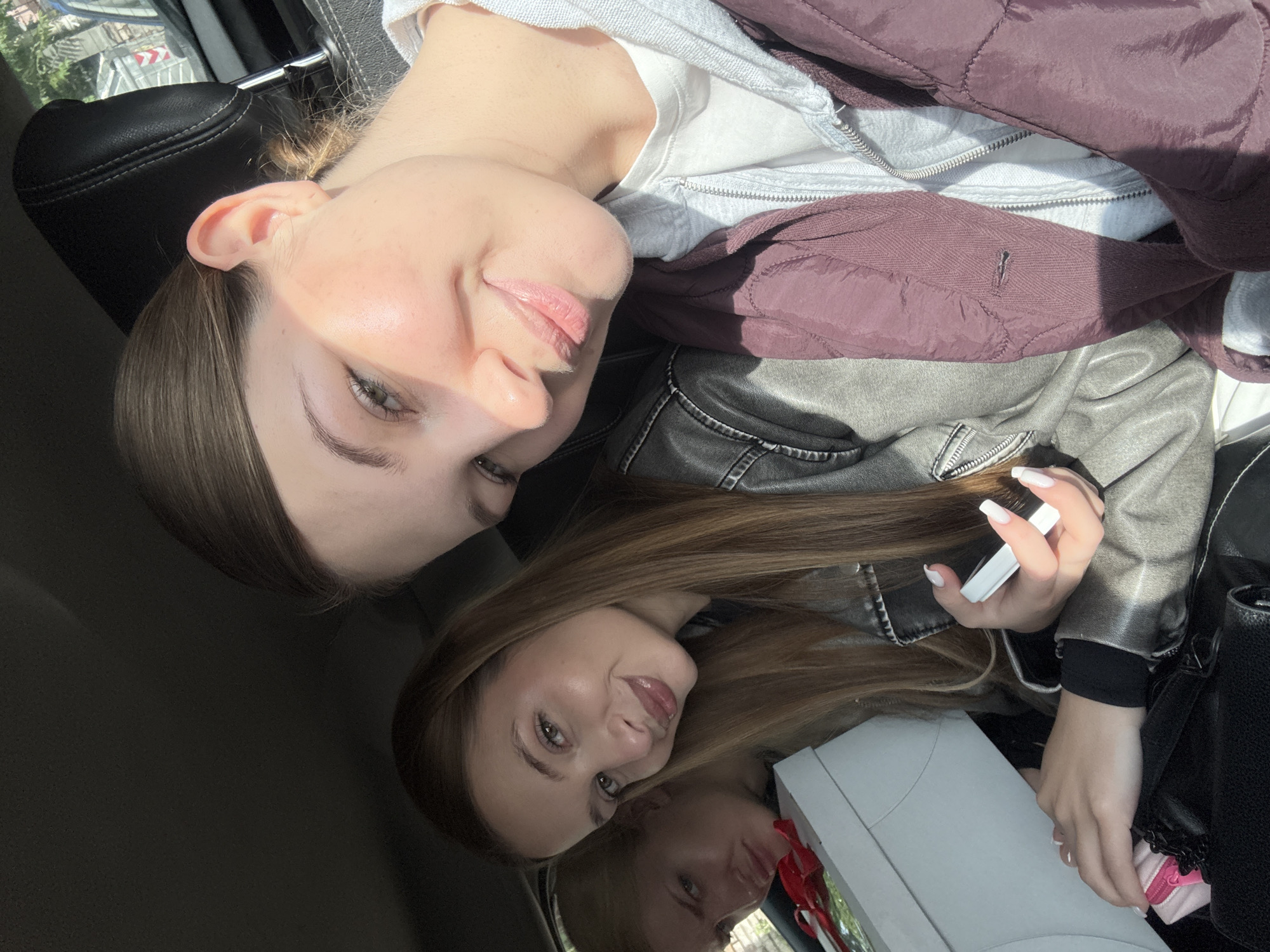 Two young women are inside a car, possibly moving based on the blurry background. The woman on the top left of the frame has short brown hair. She is wearing a white t-shirt, gray zipped hoodie, and a maroon puffy vest. The other woman on the bottom right has long brown hair. She is wearing a black shirt with a gray leather jacket. She is holding a cell phone with long white manicured nails. To her left, a large white gift box is visible. The background outside the car is blurry. The image appears to have been taken during daylight hours in Vozdovac Urban Municipality, Serbia.