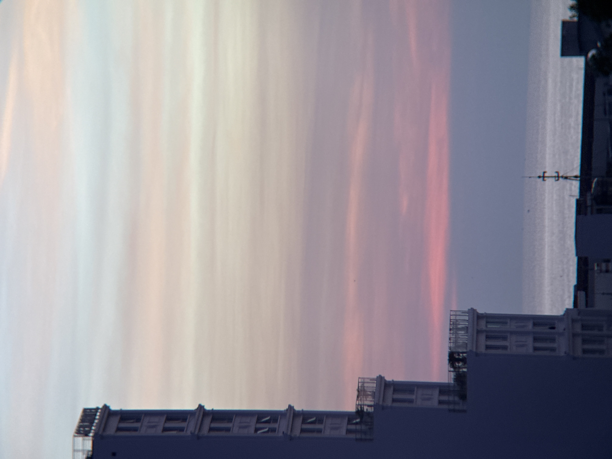 A high-angle, medium shot captures a segment of a building in Nha Trang, Vietnam, at dusk. The building, rendered in a deep blue hue, dominates the left side of the frame, its vertical lines extending upwards. Several stories are visible, featuring rows of rectangular windows and ornate balconies. The sky above is a gradient of soft pastel colors, transitioning from pale yellow and pink to a muted lavender, indicating the setting sun. A sliver of the ocean is visible on the right, its surface reflecting the muted sky. The far horizon is marked by a line of buildings and a prominent telecommunications tower. The scene is devoid of any visible people or animals.