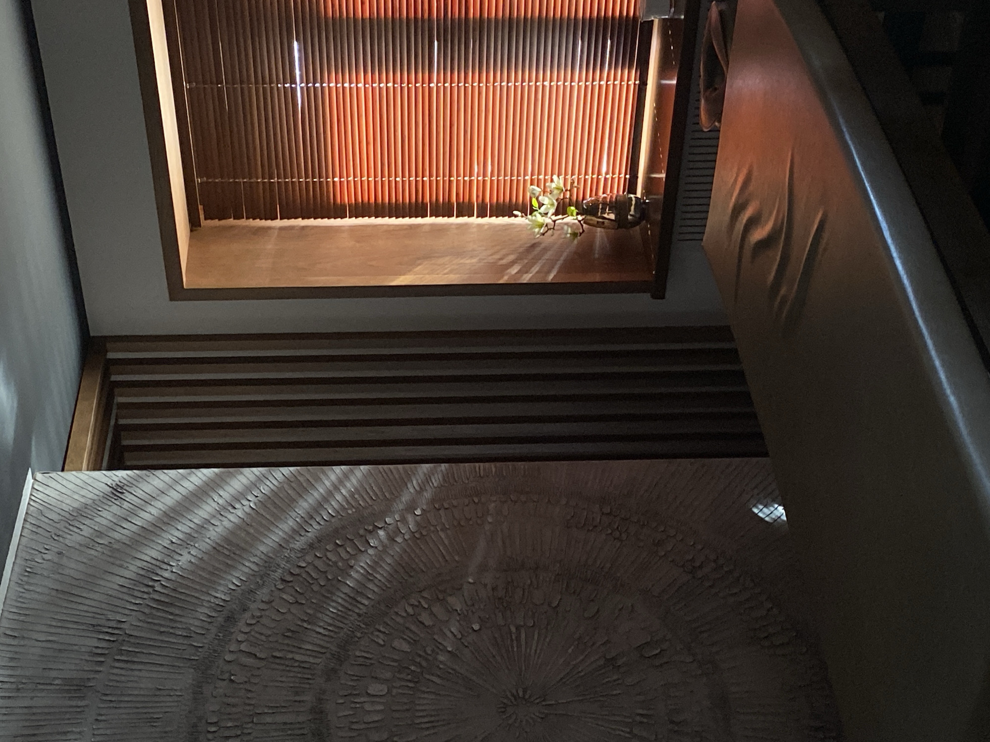 The image depicts an interior shot from an elevated perspective, featuring a variety of surfaces and textures. A window with thin, vertical slats allows light to stream through, creating contrasting light and shadow. On the ledge below the window sits a small plant or floral arrangement. To the right is a smooth, light-colored surface that appears to be a cushion or padding. Below the window is a section with horizontal wooden slats. The bottom portion of the frame showcases a large, circular pattern with a rough, textured surface, possibly a table or decorative art piece. The environment is dimly lit, contributing to a somber or contemplative mood. There is no indication of the location being Votkinsk, Russia.
