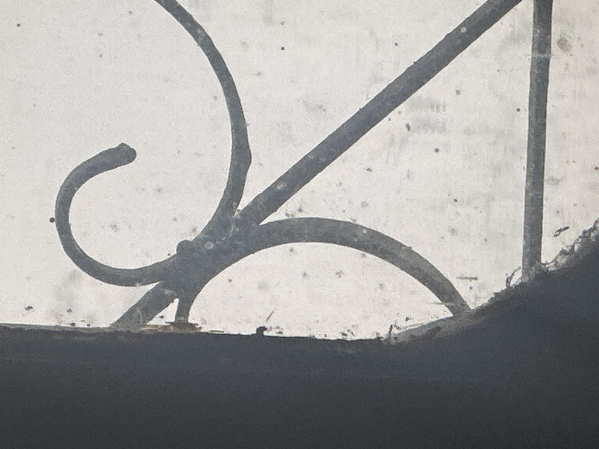 The image shows a close-up view of a decorative metal grate, likely part of a window or balcony railing. The grate is made of dark, wrought iron, with curved and linear elements. It appears to be aged, with some speckling and texture on the metal surface. The background is a muted, light color, possibly a wall or sky, with small, dark specks scattered throughout, which could be dust or debris. The left side of the image features a dark, vertical element, which seems to be a structural beam or frame, possibly covered in a rough material. The overall impression is one of an outdoor architectural detail, possibly in an older building. This scene is located in the city of Bhagsu, India.