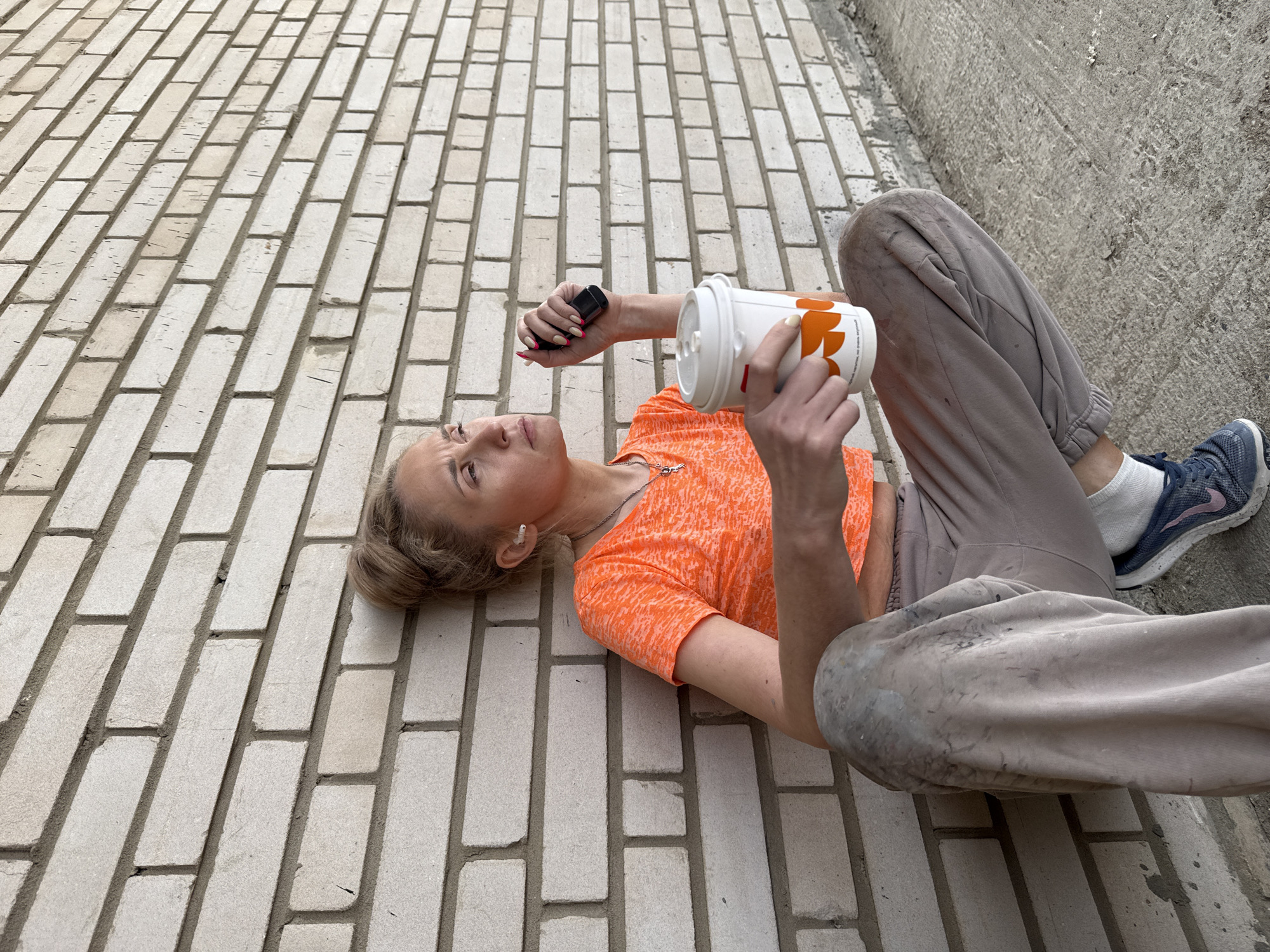 A woman lies on a brick walkway in Chaikovsky, Russia, next to a concrete curb. She wears an orange shirt and light brown sweatpants with paint stains. On her feet are blue running shoes and white socks. She holds a black electronic device in one hand and a white paper cup with orange floral designs in the other.