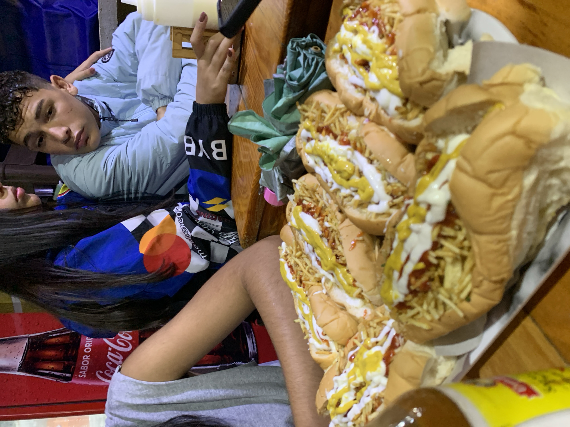 In Santa Ana, Venezuela, a group of young people are gathered around a table laden with hot dogs topped with french fries, ketchup, and mayonnaise. The scene appears to be a casual dining setting, possibly a street food vendor or a local eatery. A red Coca-Cola bottle is visible in the background, suggesting a brand preference. One of the individuals, a young man, is looking away from the camera with a slightly pensive expression. Another person, a young woman, has her arm around him, indicating a close relationship. The overall atmosphere is relaxed and convivial, with the focus on the shared meal.