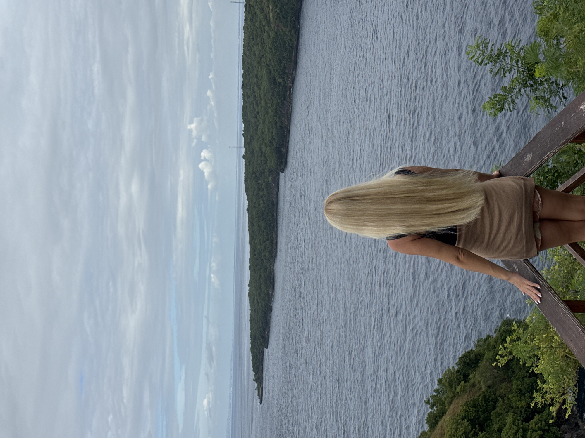 A woman with long blonde hair stands on a wooden lookout point, her back to the viewer. She is wearing a beige dress or cover-up. In the background, there is a wide body of water, with a long strip of land covered in dense green trees on the opposite shore. Above, the sky is overcast with grey clouds. The vegetation around the lookout point is lush and green. The scene is set in Nusa Penida, Indonesia.