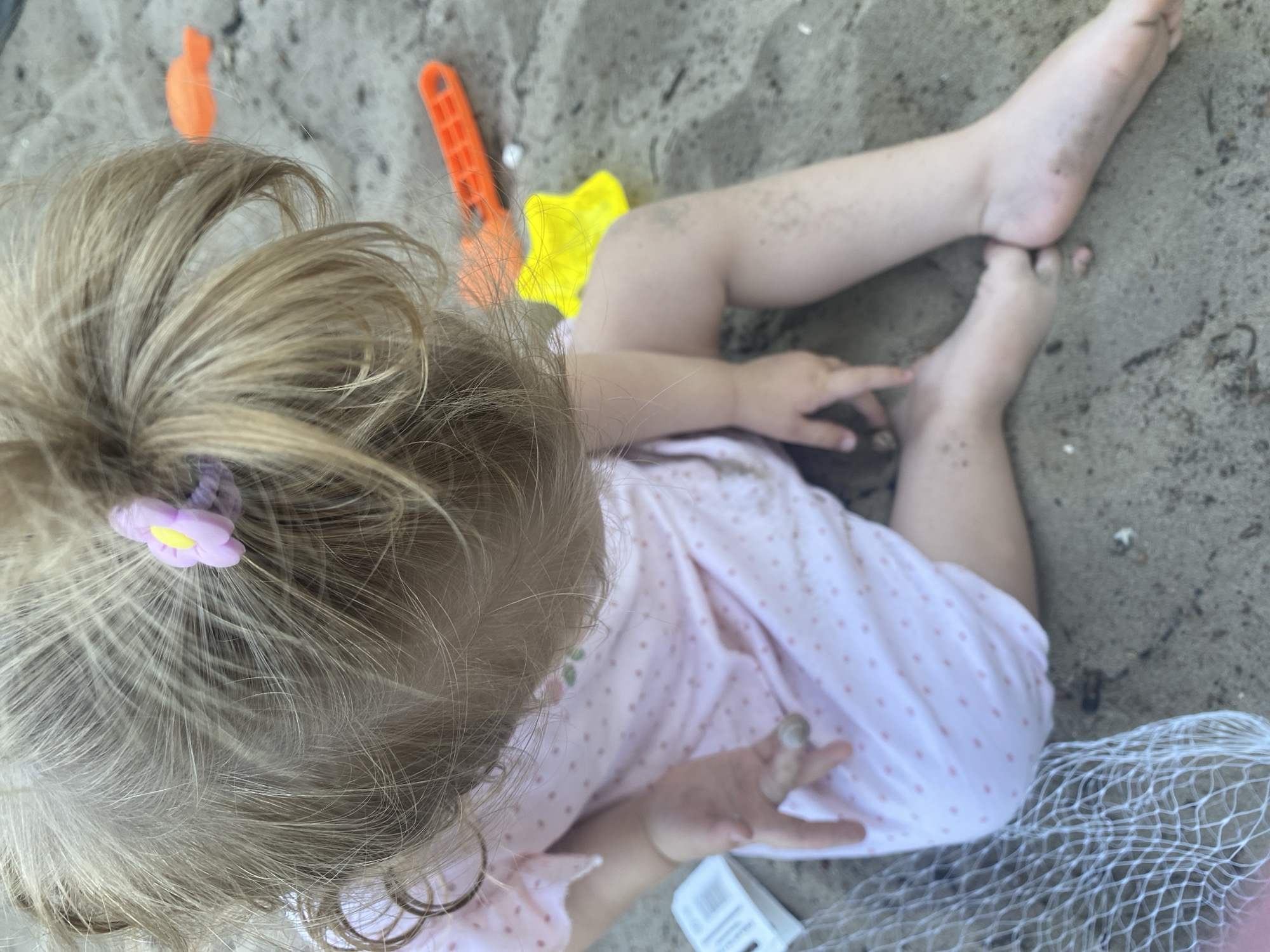 The image depicts a close-up, overhead view of a young child with blonde hair, wearing a pink polka-dot dress, sitting on sand. The child's hair is tied up with a small purple and yellow flower clip. Sand is visible on the child's legs and feet, and on their hands. In the background, there are several brightly colored children's sand toys, including an orange shovel and a yellow star-shaped mold. A white mesh bag is also present in the lower left of the frame. The scene appears to be outdoors, likely in a sandy area such as a playground or beach. There is no specific indicator of location within Dnipro, Ukraine.