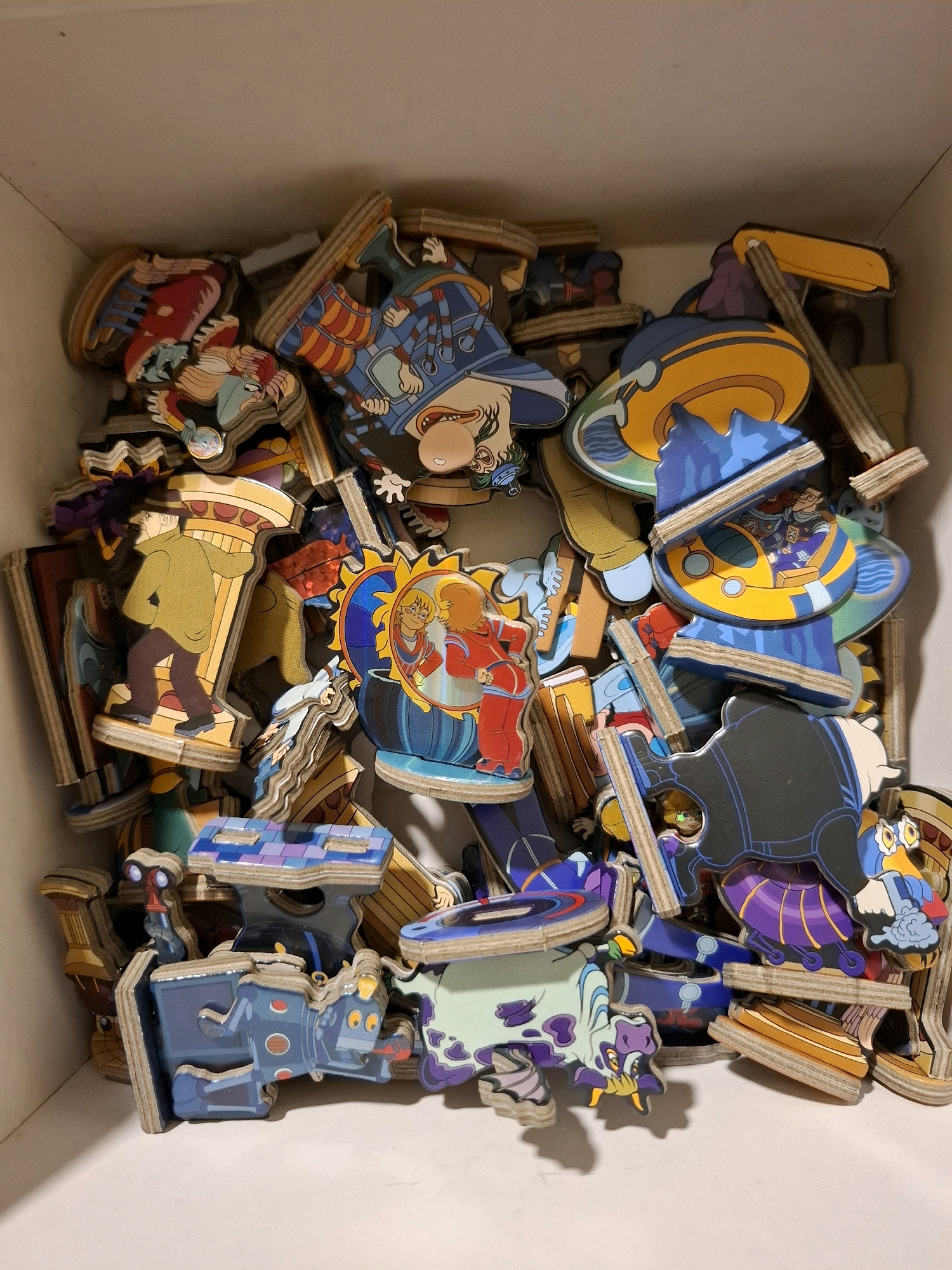 The image shows a top-down view of a box filled with scattered, wooden puzzle pieces featuring colorful cartoon characters and objects. The puzzle pieces are various shapes and sizes, and some are layered on top of each other. The box appears to be made of cardboard and is white on the inside and outside. The lighting is even, suggesting an indoor setting with overhead illumination. There is no indication of the location being in Tyumen, Russia; it is a generic indoor scene. The puzzle pieces depict animated figures in diverse costumes, including what appear to be royalty, fantastical creatures, and characters with steampunk-inspired attire. Some pieces show elements of vehicles or machinery. The overall impression is one of a disassembled puzzle or a collection of themed puzzle pieces stored within a container.