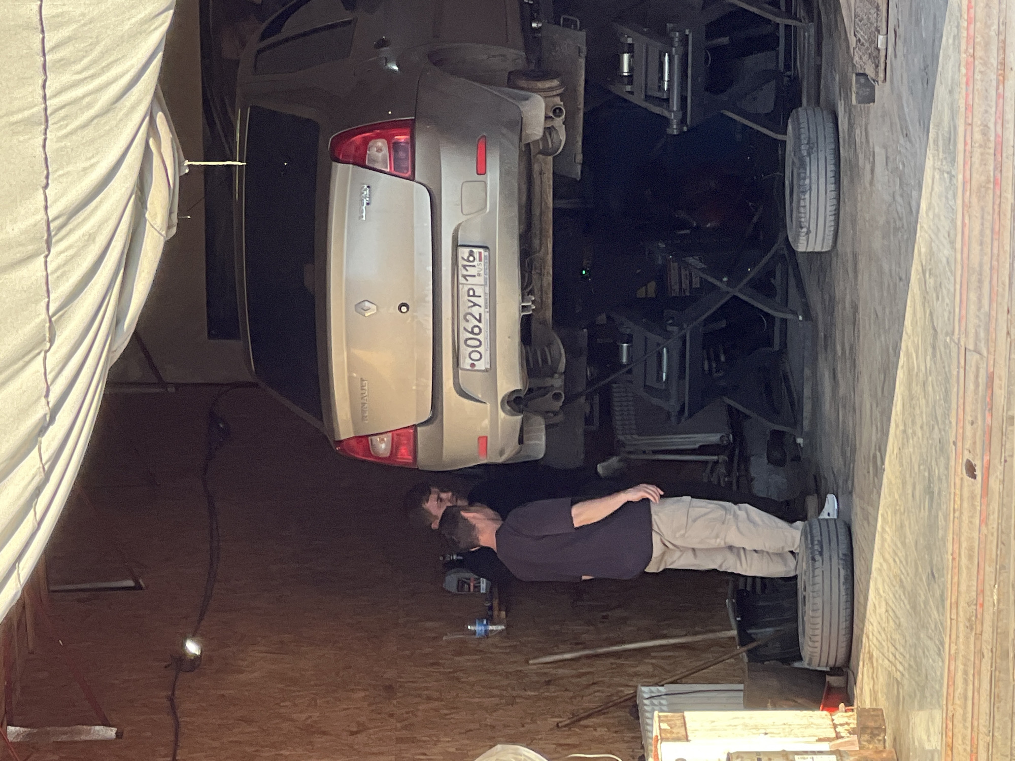 Two men are inspecting the underside of a silver Renault Logan car, elevated on a mechanical lift in a garage. The car's Russian license plate reads "O062YP 116", indicating registration in the Republic of Tatarstan, likely Kazan, Russia. One tire is positioned on the ground. A white sheet or tarp is visible on the left.
