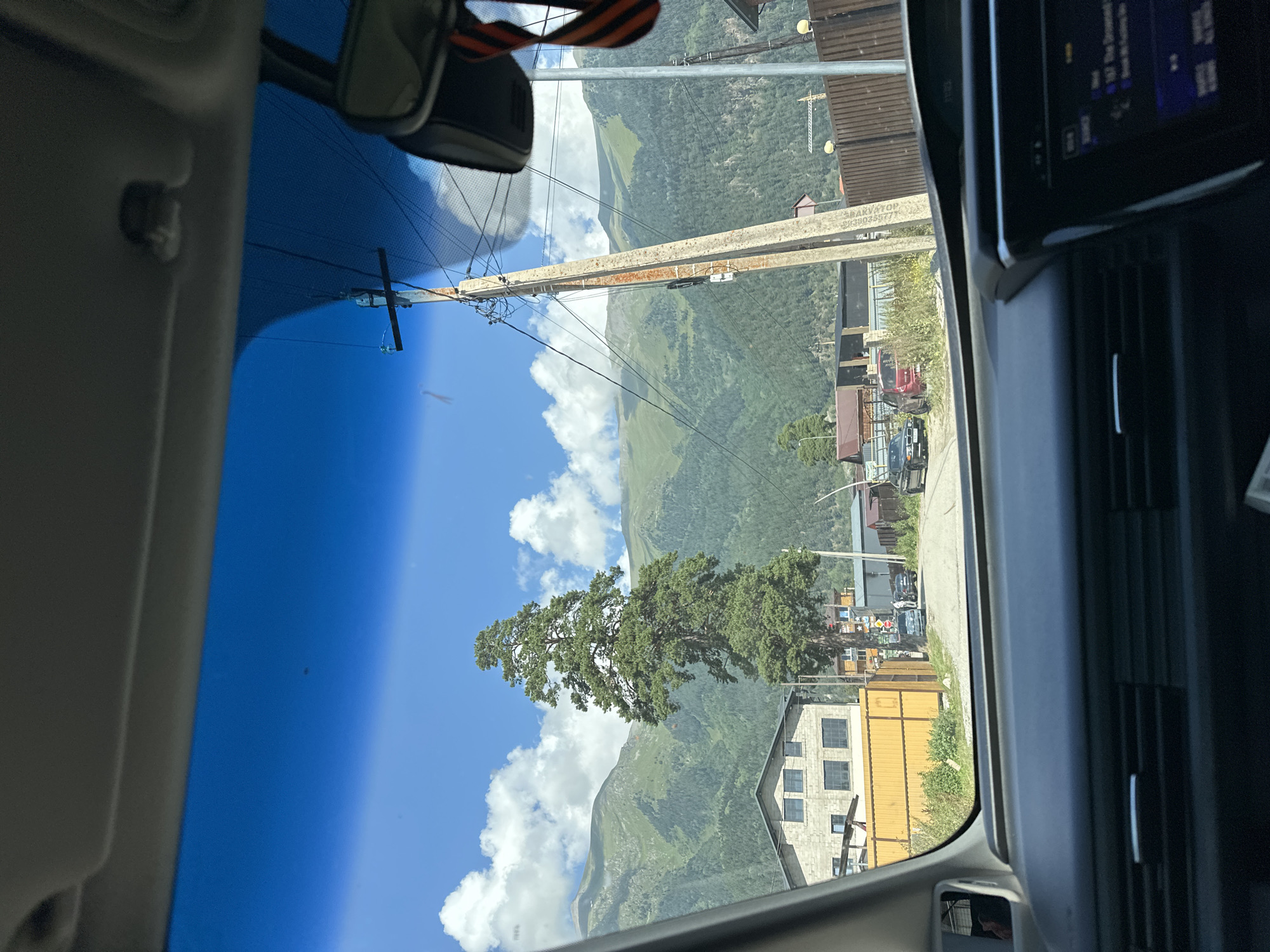 This is a point-of-view shot from inside a vehicle looking through the windshield towards a street scene in the city of Arkhyz, Russia. In the foreground, the dark gray dashboard of the car is visible, with a portion of the infotainment screen showing some text. The windshield itself is slightly tinted blue.
Outside the vehicle, a paved road leads into the distance. On the left side of the road, there is a large, light-colored, multi-story building with dark windows, preceded by a tall yellow fence. A very tall, mature evergreen tree stands prominently in front of this building. To the right of the road, there is a wooden fence and various smaller buildings and structures. A tall, weathered utility pole with numerous wires stretches vertically along the right side of the frame.
Further down the road, a few cars are parked or in motion. In the background, the scene is dominated by rolling green mountains covered in dense forest, with patches of clouds in a clear blue sky. The overall impression is a rural mountainous landscape with signs of human habitation.