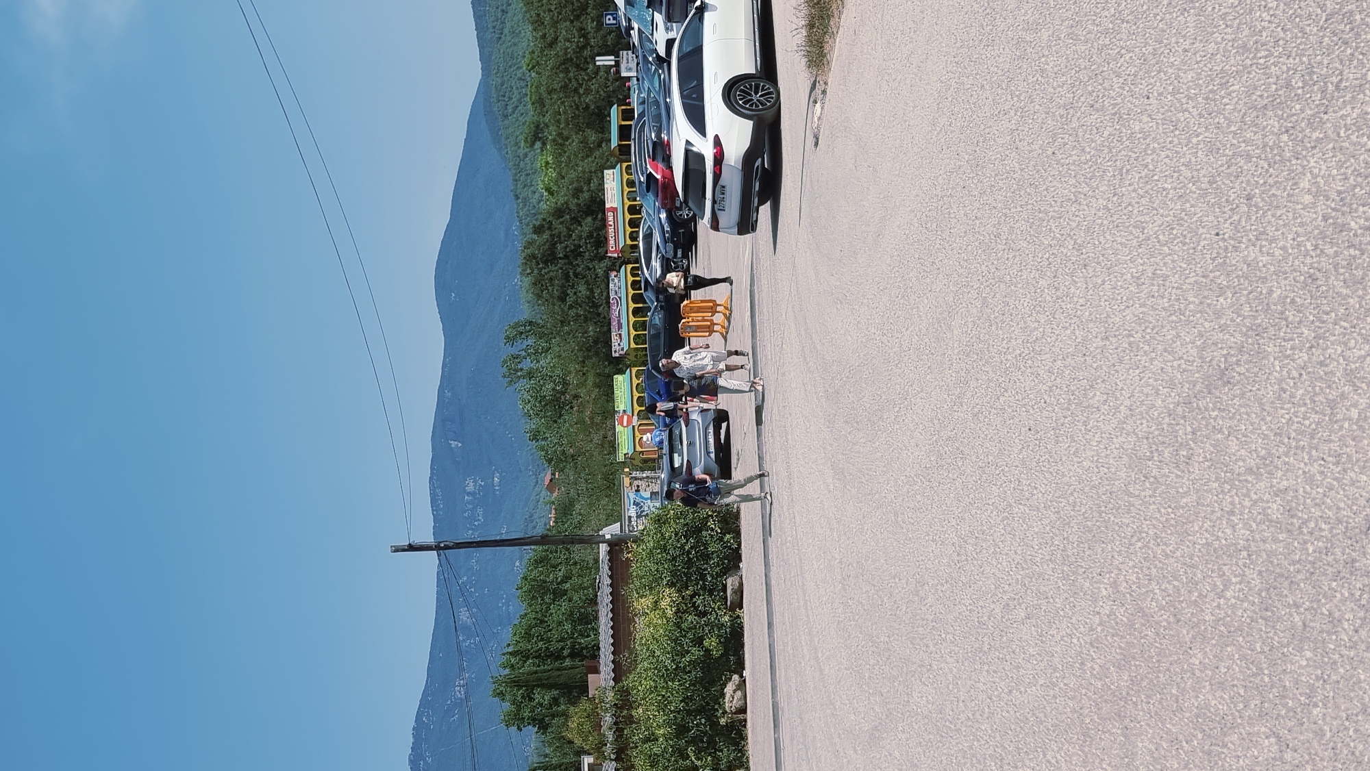 The image shows a parking lot in Besalú, Spain, with a backdrop of lush green trees and distant blue mountains under a clear sky. Several cars are parked in the lot, including a white SUV in the foreground. A group of people are walking across the parking lot, some heading towards the cars and others walking away. In the background, a train-like attraction with colorful carriages is visible, with the text "CIRCUSLAND" on one of the carriages. The overall scene depicts a sunny day at an outdoor venue.