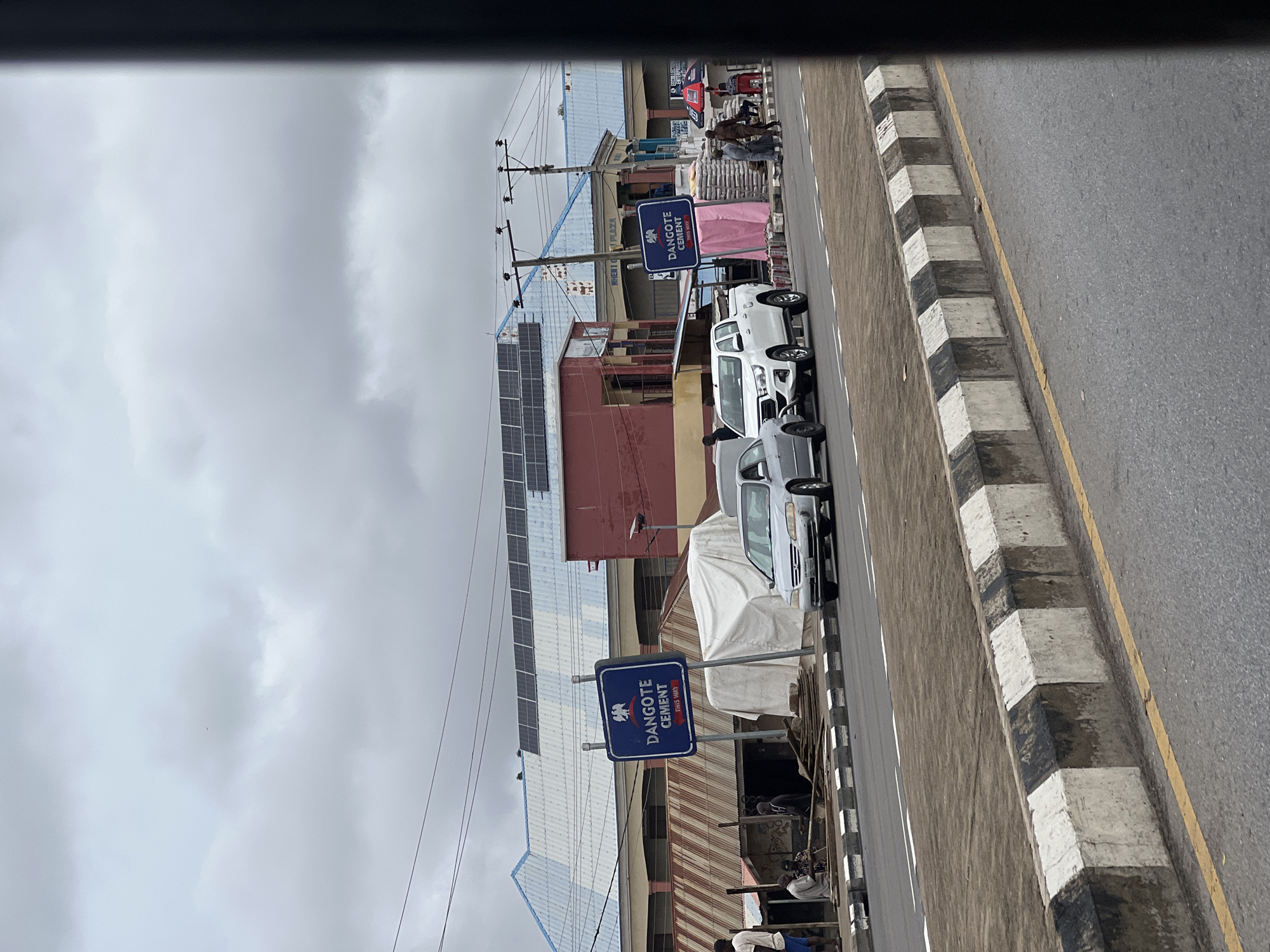 The image depicts a street scene in Jalingo, Nigeria. In the foreground, a gray sedan and a white pickup truck are parked on the right side of the road. The road is bordered by a sidewalk with a concrete barrier painted black and white. Behind the vehicles, there are several buildings. One building has a blue and white corrugated metal roof with solar panels installed. A blue sign with the text "DANGOTE CEMENT THIS WAY" is visible. To the right of this sign, there are stacks of bagged cement. Several people are seen standing and walking near the buildings and sidewalk. The sky is overcast with gray clouds. The overall environment appears to be a commercial area within the city.