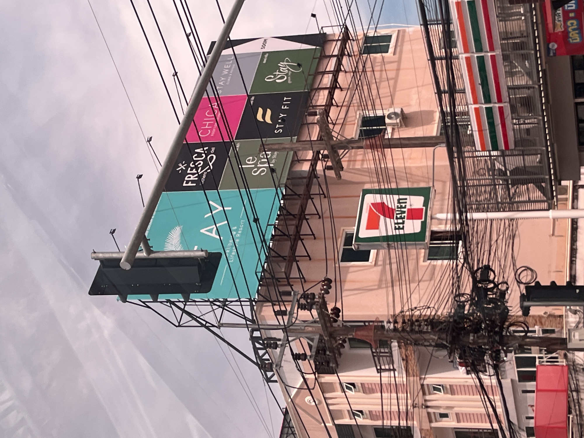 The image shows a street scene in Rawai, Thailand, characterized by a complex network of overhead electrical wires and a multi-story building adorned with various signs. A prominent billboard structure displays multiple advertisements, including "AY," "FRESCA," "CHICA," "STAY WELL," "le spa," "STAY FIT," and "Stay." Below the billboard, a building with a pink facade has windows and balconies. A "7-Eleven" sign is visible on the side of the building, and a part of the distinctive "7-Eleven" store awning with its red, green, and orange stripes is also present. A traffic light is partially visible in the lower left corner. The sky above is overcast with streaks of clouds.