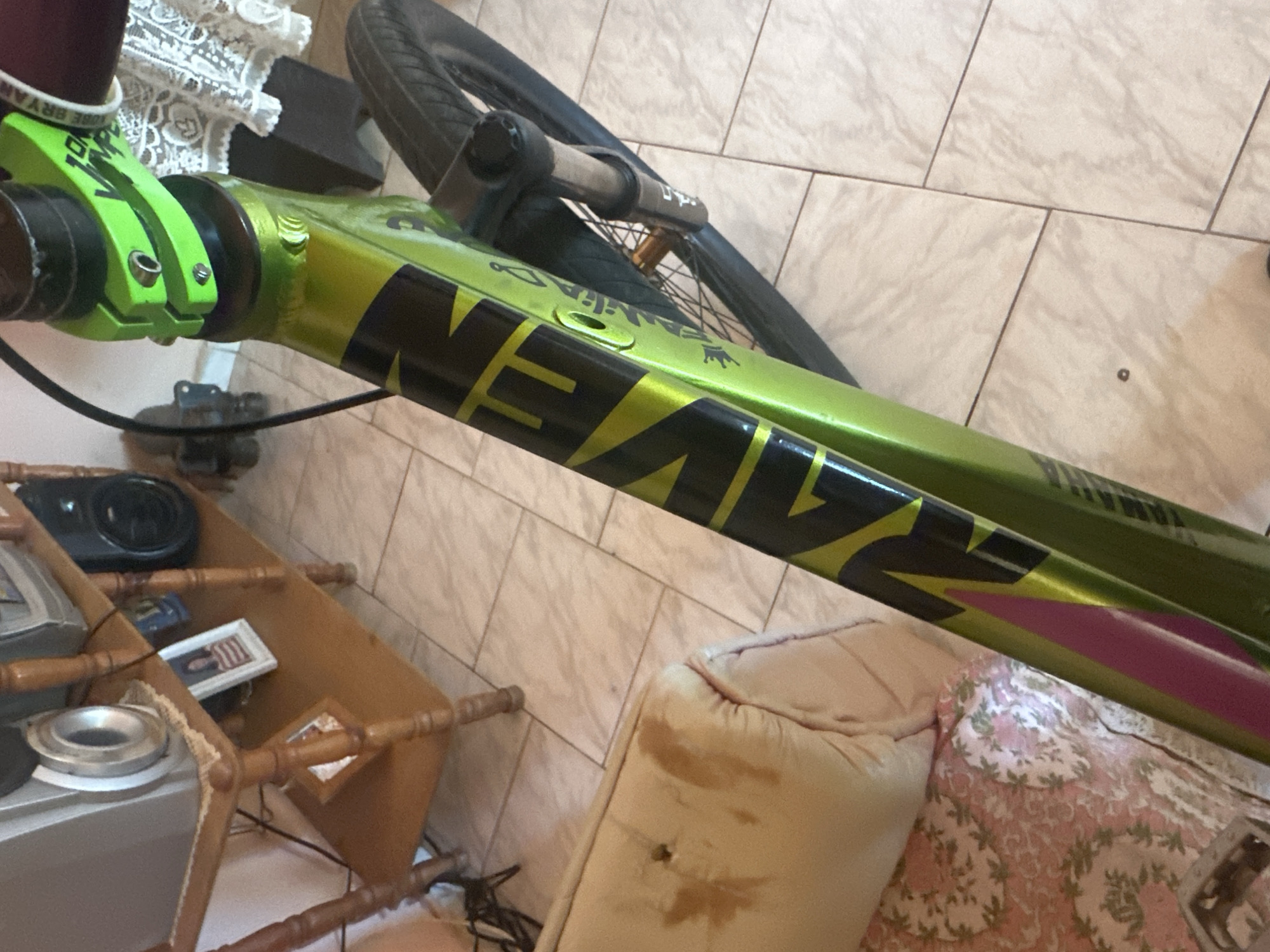 The image shows a close-up of a green bicycle frame with black and yellow lettering that appears to spell "RAVEN" and "YAMAHA". The bicycle is partially leaning against a light-colored couch. In the background, there is a wooden shelf unit with a speaker and framed photos. The scene is indoors, likely in a residential setting. The floor is tiled. The city of El Maco, Venezuela is not depicted in the image.