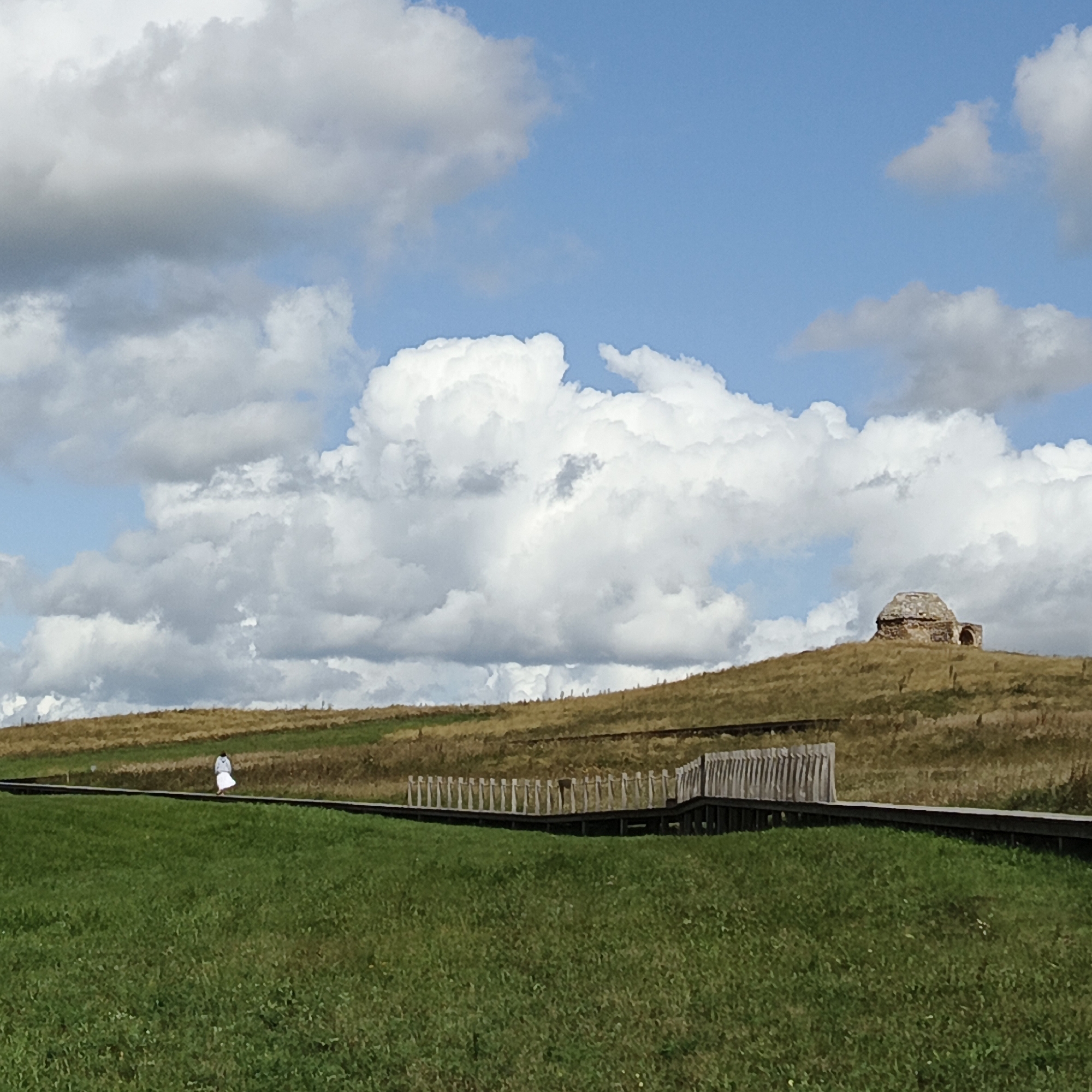 The image depicts a landscape scene under a partly cloudy sky, with a prominent dome-shaped structure atop a grassy hill in the background. A wooden walkway extends from the foreground towards the hill. In the mid-ground, a solitary figure wearing a white dress or robe walks along the walkway. The hill is covered in dry, yellow-brown grass, with some green patches visible lower down. The sky is blue with large, white cumulus clouds. The dome structure appears to be made of stone and has an arched opening. The wooden walkway is elevated and has a rustic wooden fence bordering one side. The foreground consists of a lush green grassy field. This scene is located in Verkhnie Termy, Russia, specifically at the site of the ancient settlement of Old Izborsk, known for its historical ruins and natural beauty.