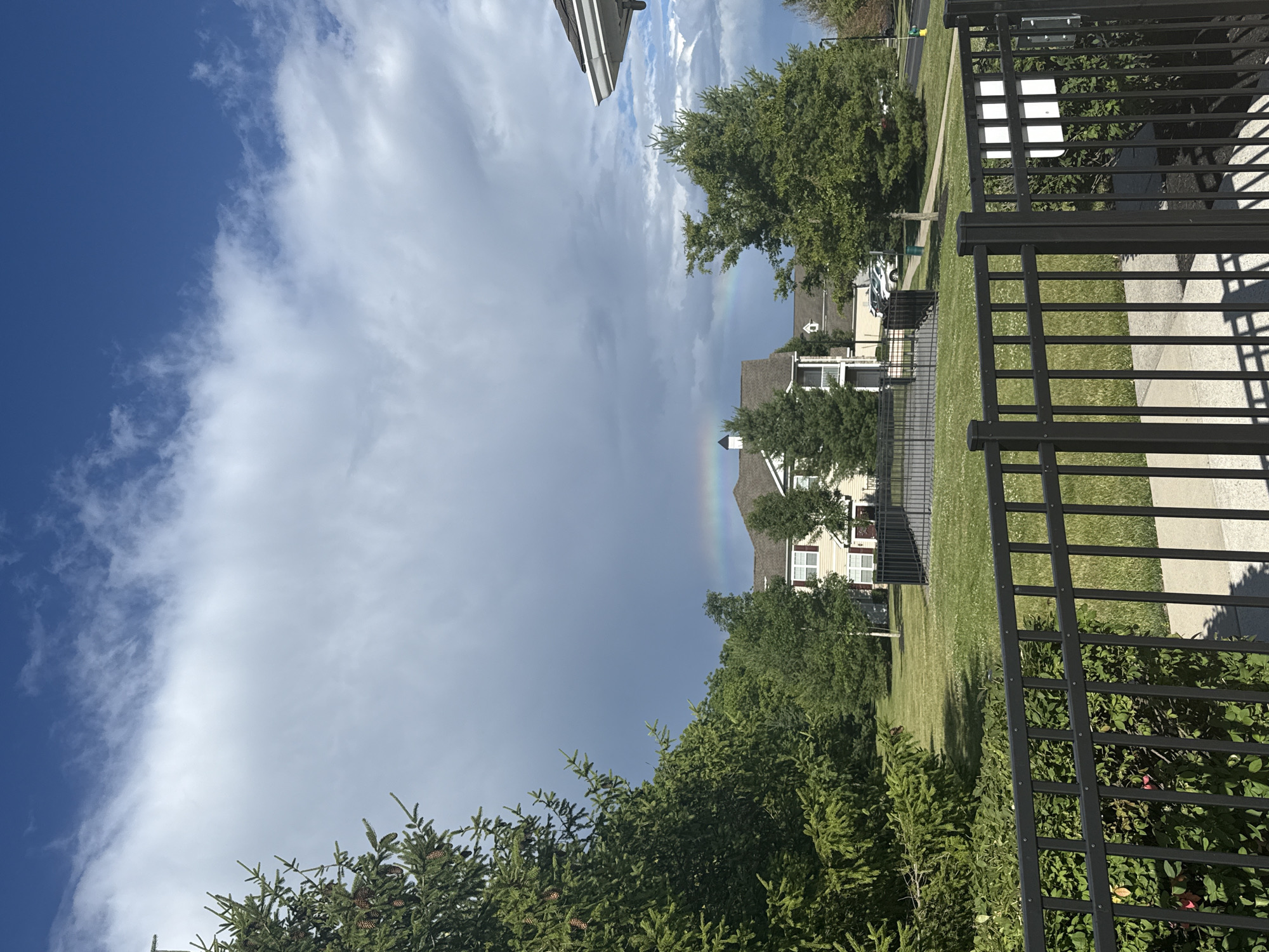 The image shows a residential area with a faint rainbow visible in the sky. In the foreground, there is a black metal fence. Beyond the fence, a manicured lawn is visible, with trees and a two-story building in the background. The building appears to be a residential complex with multiple windows and a light-colored exterior. The sky is a mixture of blue sky and dark clouds, suggesting recent or impending rain. The scene is set during daylight hours.