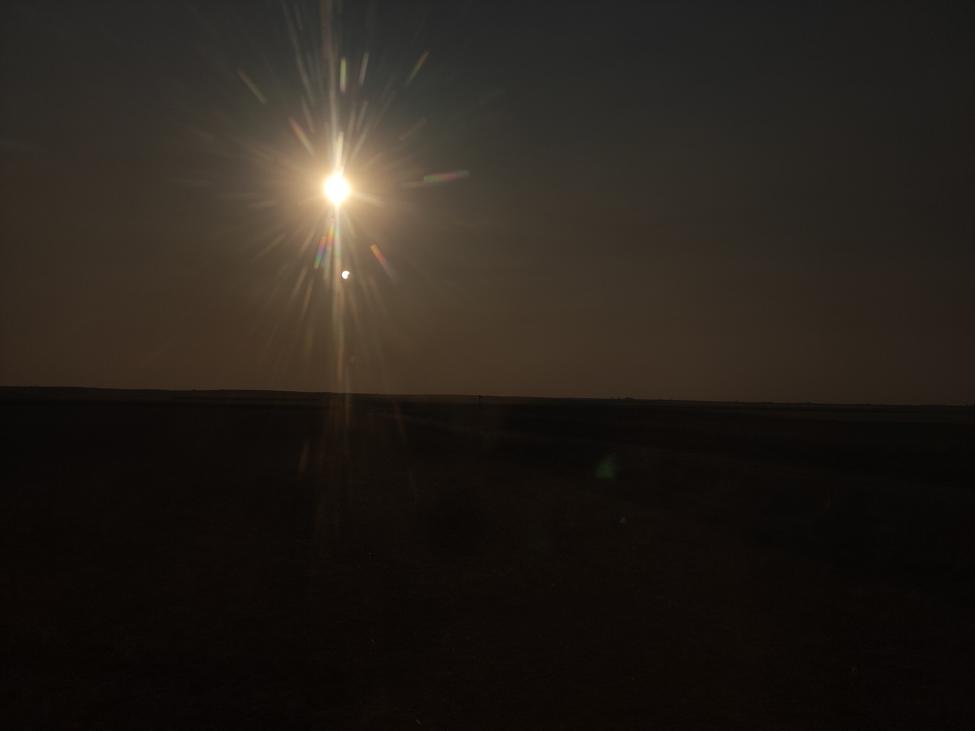 The image shows a wide, flat landscape under a dark, hazy sky. The sun is prominently featured in the upper left quadrant, emitting bright rays and lens flare. A small, darker orb, likely the moon, is visible just to the right of the sun, indicating a solar eclipse. The horizon line is low and indistinct, separating the dark sky from the equally dark ground, which appears to be a field or open terrain. There are no visible structures or people in the landscape. The overall impression is one of a desolate, open space during a celestial event.