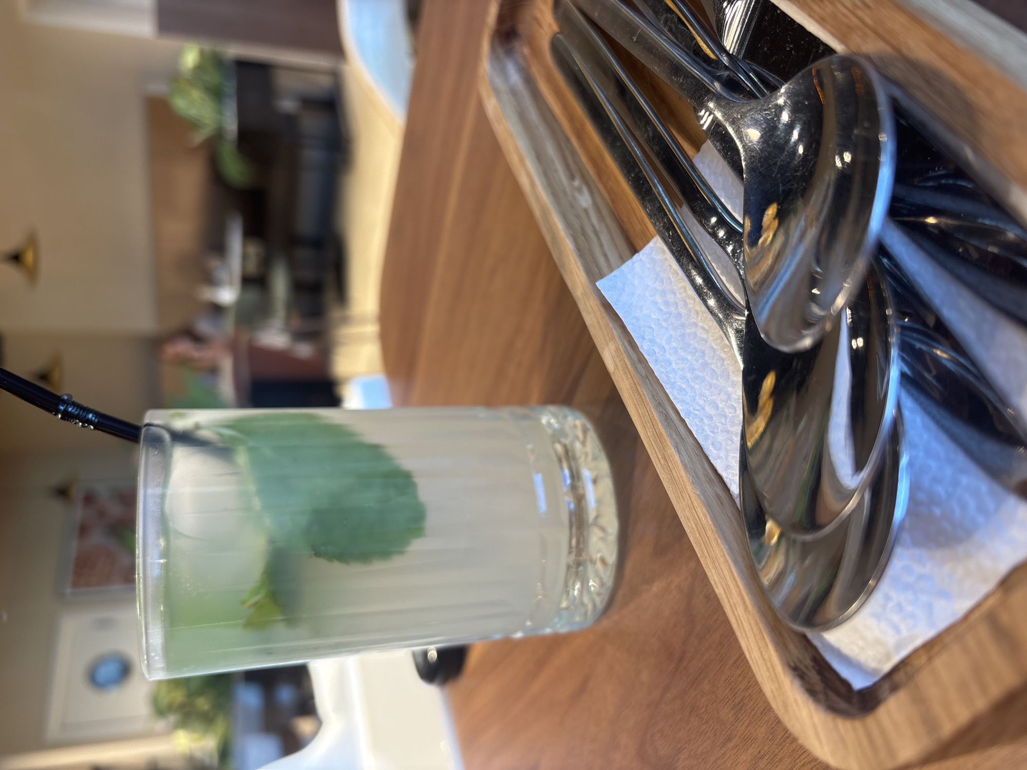 A tall glass of a pale yellow beverage with mint leaves and ice cubes is positioned on a wooden table. To the right of the glass, a wooden tray holds several shiny metal spoons, with a white napkin underneath them. The background is blurred, showing the interior of what appears to be a cafe or restaurant, with seating areas, tables, and a few people seated in the distance. The lighting suggests an indoor setting, likely during daytime. The city of Naberezhnye Chelny, Russia is not identifiable from the visual information presented in the image.