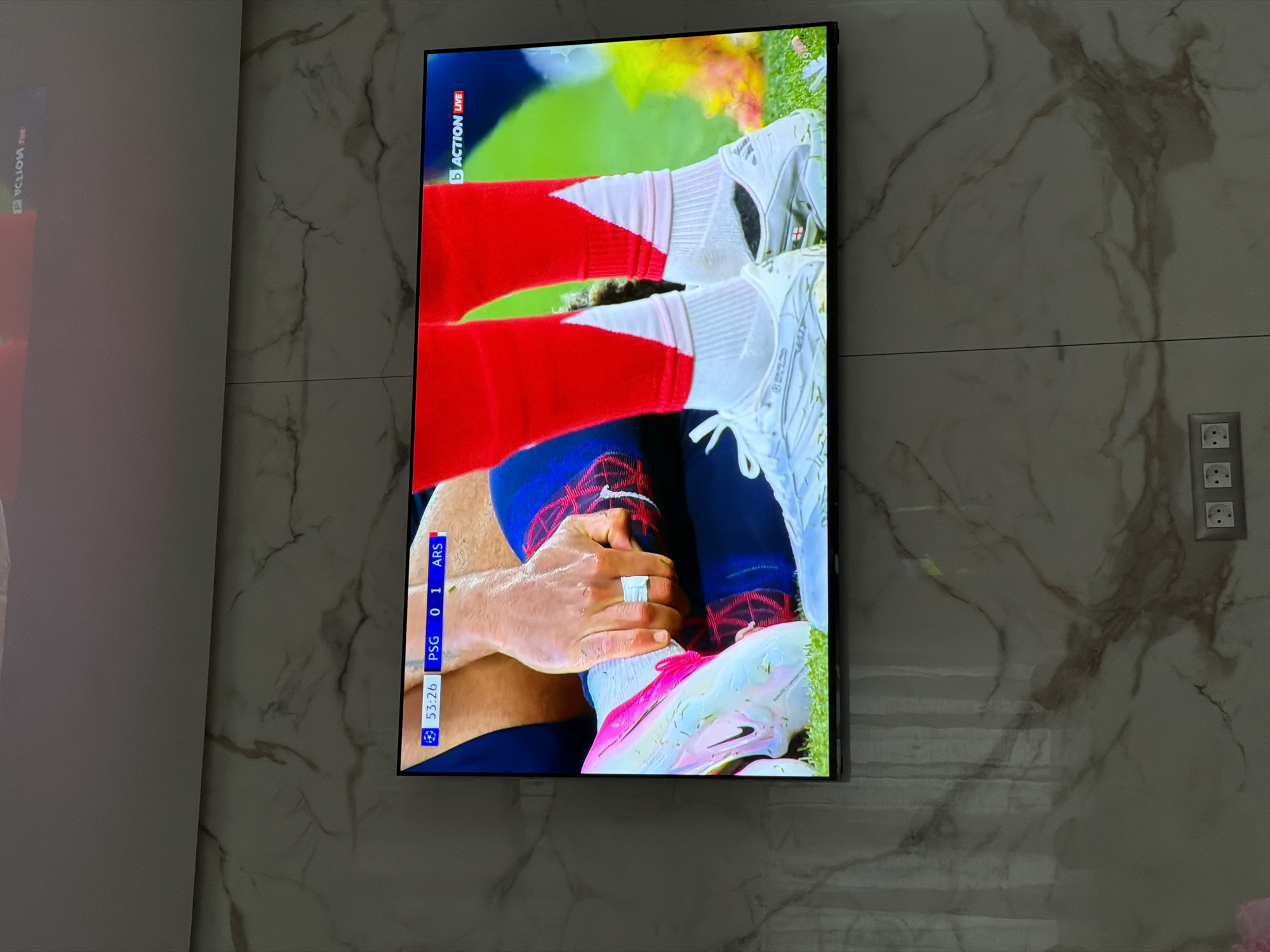 A television displays a soccer game with the score PSG 0 - 1 ARS, time 53:26. Players are shown with red socks and white and pink shoes. An individual's hand is visible, wrapped in tape, likely indicating an injury. The television is mounted on a marble-patterned wall in an interior setting. An electrical outlet is visible on the right. The location is unknown.