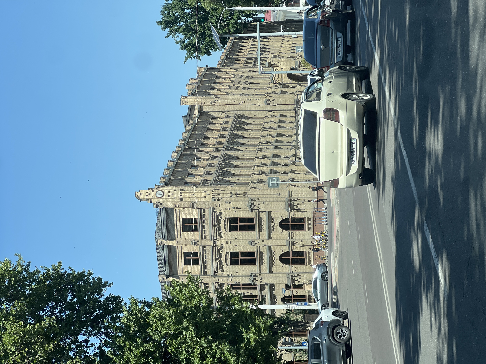 In Tashkent, Uzbekistan, on a clear, sunny day, a long, light-brown brick building with an ornate facade stands prominently on the left side of a paved city street. The building features numerous rectangular and arched windows, decorative brickwork, and a clock tower on the right side. A large, green tree with dense foliage partially obscures the left side of the building.
Several vehicles are present on the street, with a light-colored sedan in the foreground and a dark-colored sedan behind it, both facing away from the viewer. Other cars are visible further down the road. On the sidewalk in front of the building, a group of people are seen, some with bicycles and electric scooters. Traffic lights and street lamps are also present. The sky is a bright, clear blue with a few faint specks, possibly birds or dust. The scene appears to be a typical urban streetscape with architectural and traffic elements.