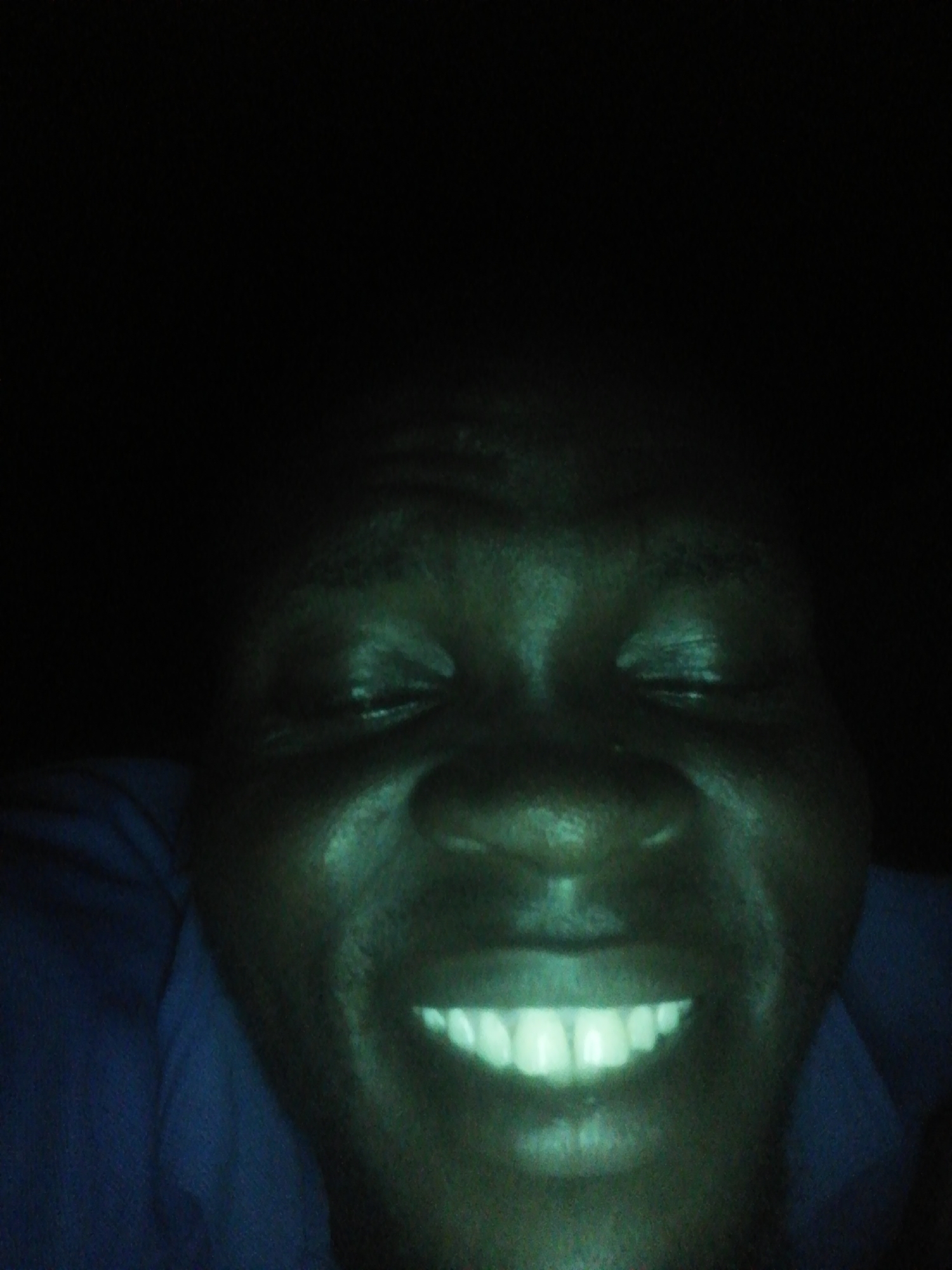 A close-up photograph of a dark-skinned person's face, focusing on the area from the chest up, is set against a black background. The person is smiling, revealing a row of teeth that stand out due to their lighter color in contrast to the darker skin tone. The photograph appears to have been taken in low-light conditions, which affects the clarity and visibility of the subject. No specific location is discernable within the city of Unknown, Unknown.