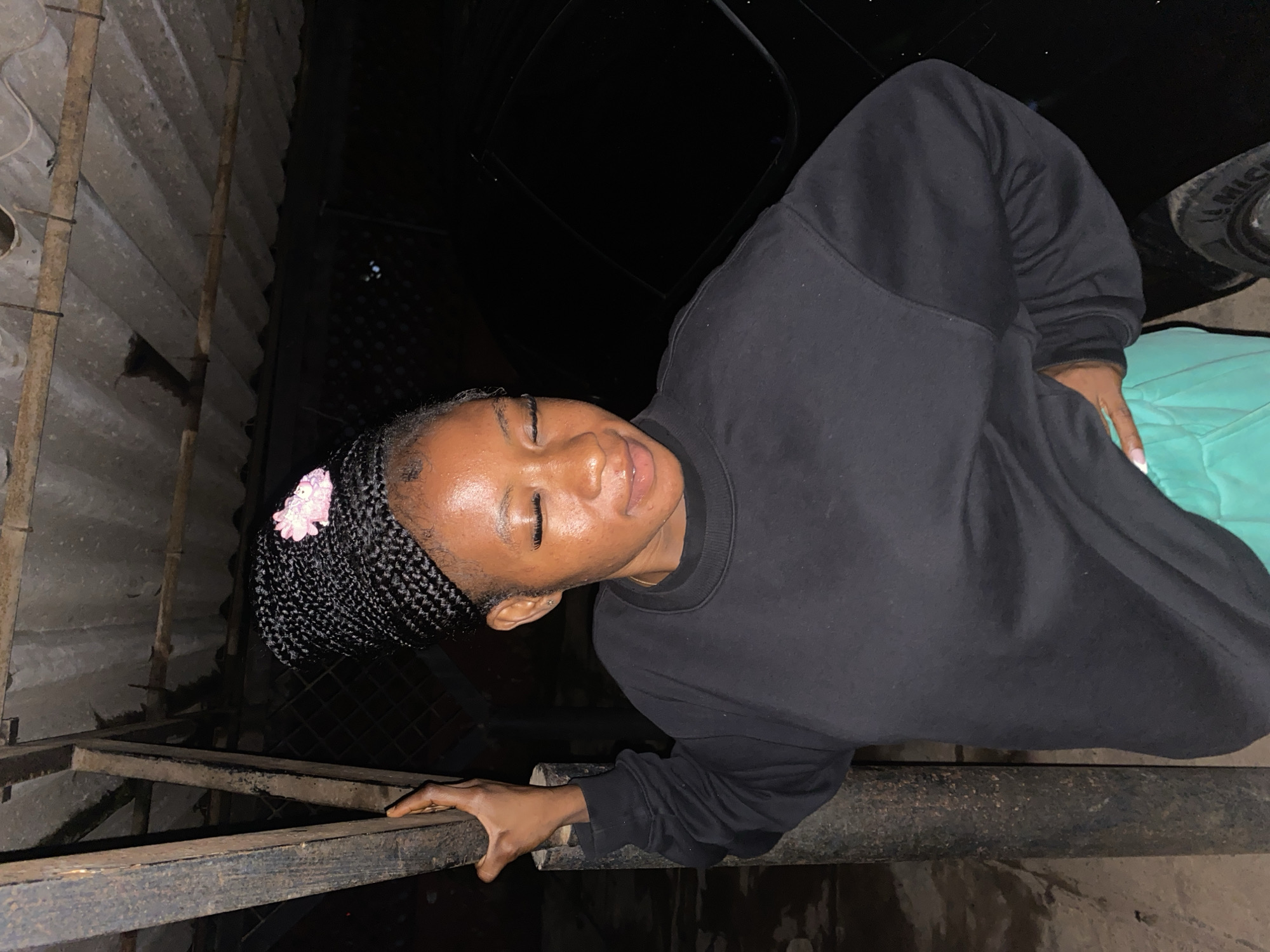 In a dimly lit outdoor setting, likely a covered outdoor area, a young woman of African descent is standing and posing for a photograph. She has braided hair styled in a high bun adorned with a small pink flower. Her eyes are closed, and she is wearing a black crew-neck sweatshirt and light green pants. Her left arm is bent, and her hand rests on a dark vertical pole. Her right hand is placed on her hip. Behind her, a dark vehicle is visible, and the ground appears to be unpaved. The overhead structure is made of corrugated roofing material supported by wooden beams. The scene's lighting suggests it is nighttime. The specific location is within the city of Jos, Nigeria.