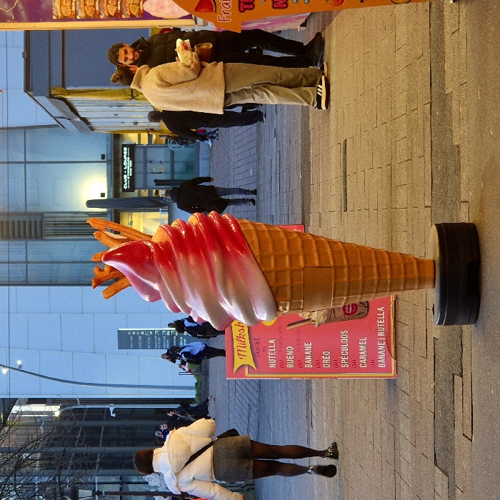 The image depicts a bustling pedestrian area within the Presqu'île André Malraux district of Strasbourg, France, at dusk or early evening.
In the foreground, a large, free-standing replica of a soft-serve ice cream cone dominates the center, featuring a textured beige waffle cone and a prominent swirl of red and white ice cream. It stands on a black circular base. To its immediate left, a pink A-frame sign displays a "Milkshake" menu in French, listing flavors such as "FRAISE," "NUTELLA," "BUENO," "BANANE," "OREO," "SPECULOOS," and "CARAMEL." A vertical sign for "Fraise" (Strawberry) is partially visible on the far right, suggesting a nearby food stall.
In the midground to the left, a person with dark hair, clad in a white padded jacket, dark shorts, and dark hosiery, walks away from the viewer, carrying a dark shoulder bag. To the right in the midground, a man and a woman are conversing near an unseen stall. The woman wears a light-colored, possibly beige, jacket and light trousers, with Adidas sneakers, holding a white container. The man wears a dark puffer jacket.
The background features a paved pedestrian walkway constructed from grey interlocking bricks. Modern glass and light-colored buildings with vertical architectural details line the area. A curved streetlamp is visible on the left. Multiple other individuals are walking throughout the background. A directory board displaying names such as "HORIZON," "ADAGE," "ARCADIA," "EURÔPA," and "STRASBOURG" is visible, corroborating the location. A "CLUB - LOUNGE" sign is discernible on a building facade further back. The scene is illuminated by a mix of ambient artificial light and residual daylight.