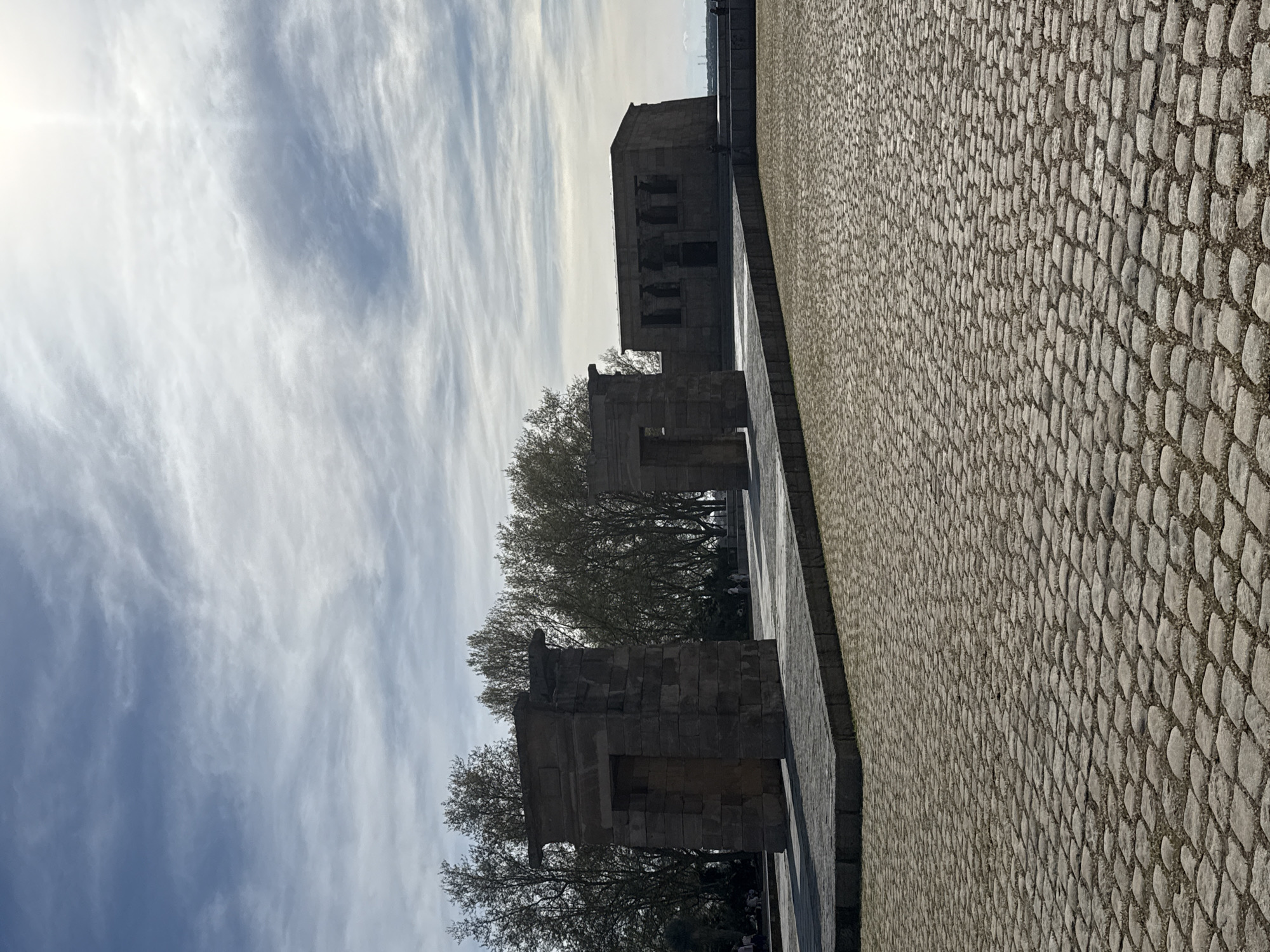 The image depicts the Temple of Debod in Madrid, Spain. The temple is constructed of weathered stone blocks and consists of several distinct structures. The foreground features a paved area composed of small, irregularly shaped stones. Behind the temple, trees are visible. The sky is light blue with wispy clouds.