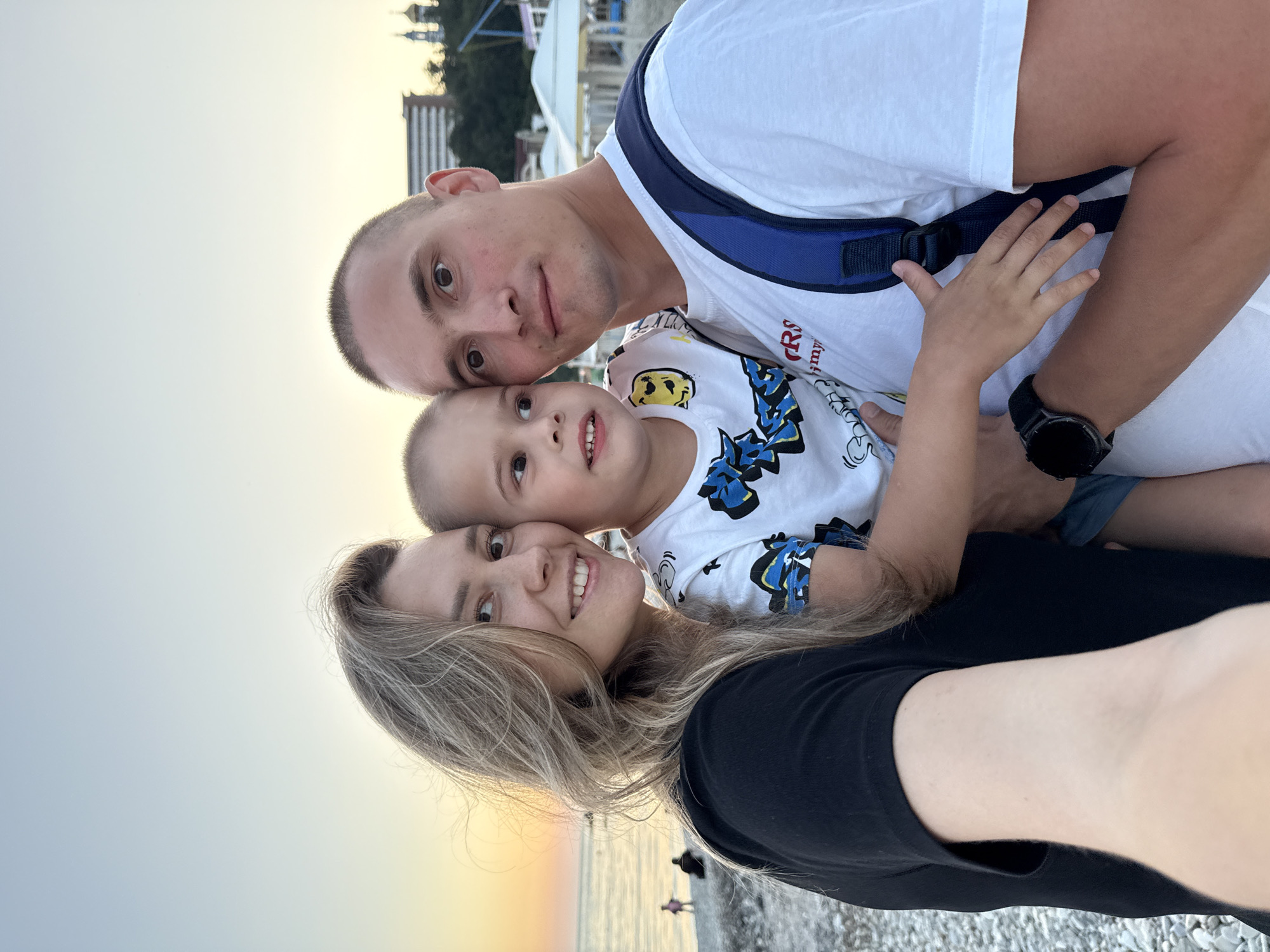 A family of three, a woman, a man, and a young boy, are taking a selfie on a pebble beach in Nebug, Russia. The sky is a soft gradient of orange and blue, suggesting sunset. In the background, the Black Sea is visible, with a few figures walking on the shore. Further inland, there are buildings and some white umbrellas on the beach. The woman is on the left, wearing a black top, and the man is on the right, wearing a white t-shirt and a blue backpack. The boy is in the middle, held by the man, wearing a white t-shirt with a graphic print. They are all looking at the camera, and the scene appears relaxed and happy.