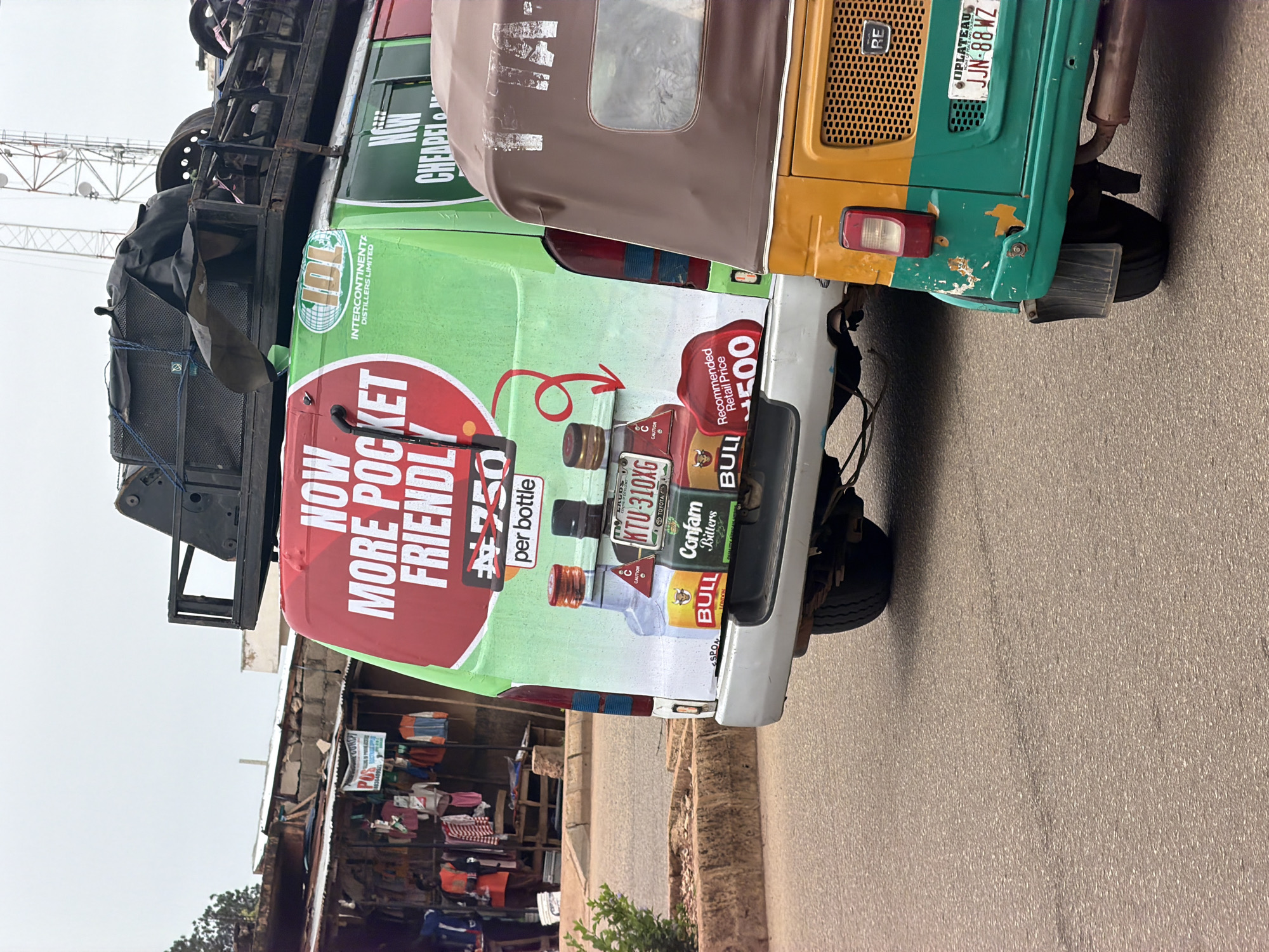 This image depicts a street scene in Bukuru, Nigeria. In the foreground, a green and yellow three-wheeled auto rickshaw is parked on the right side of the frame, facing away from the viewer. Its rear license plate reads "JJN-88WZ" and "JOS PLATEAU". Behind the auto rickshaw, the rear of a white van is visible. The van is adorned with a large, green advertisement featuring a red circle with white text that reads "NOW MORE POCKET FRIENDLY" and "#750 per bottle". Below this, there are images of three liquor bottles with labels that include "BULL", "Confam Bitters", and "Recommended Retail Price #500". The van also has a license plate with the text "KTU-31OXG". In the background, a roadside market stall with various items of clothing for sale is visible on the left. The sky is overcast.