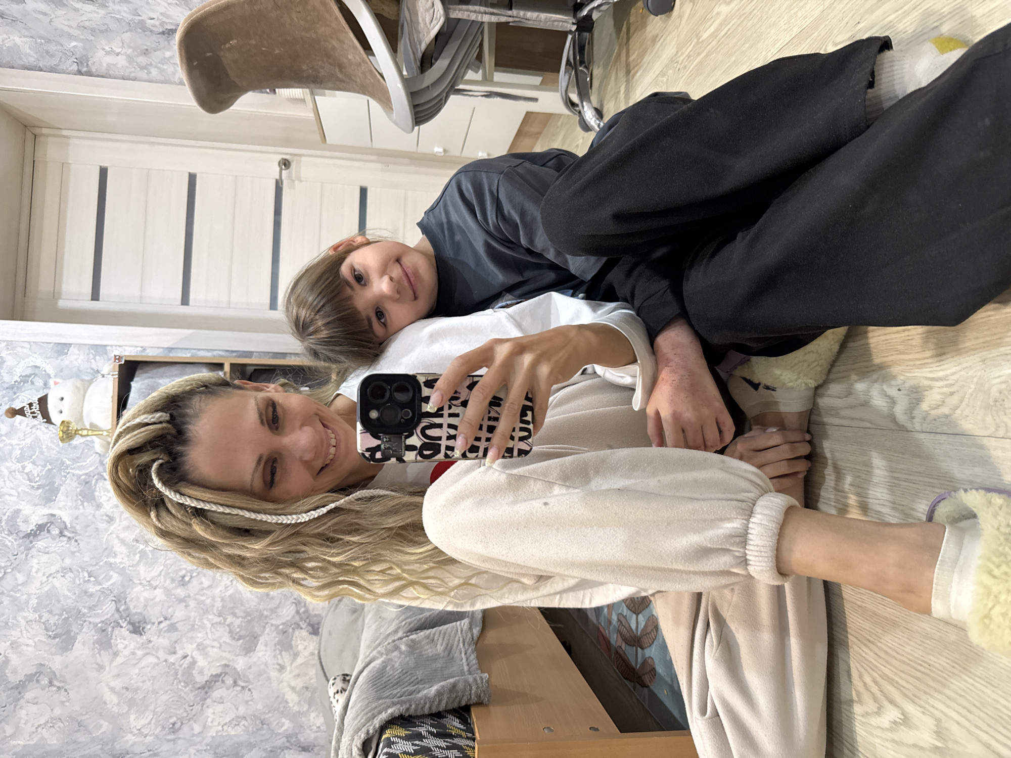 A woman with blonde braided hair is lying on the floor, holding a phone in her right hand, with her arm extending towards the camera. She is wearing a white long-sleeved shirt and light beige sweatpants. A young child with brown hair is lying on top of her, looking at the camera. The child is wearing a dark shirt and dark pants. They both appear to be taking a selfie. The floor is wooden. In the background, there is a door, a chair, a shelving unit with a stuffed animal, and a wall with a gray and white pattern. The location is likely indoors, potentially in a home. There is no information in the image to confirm it is the city of Rusanovo, Russia.
