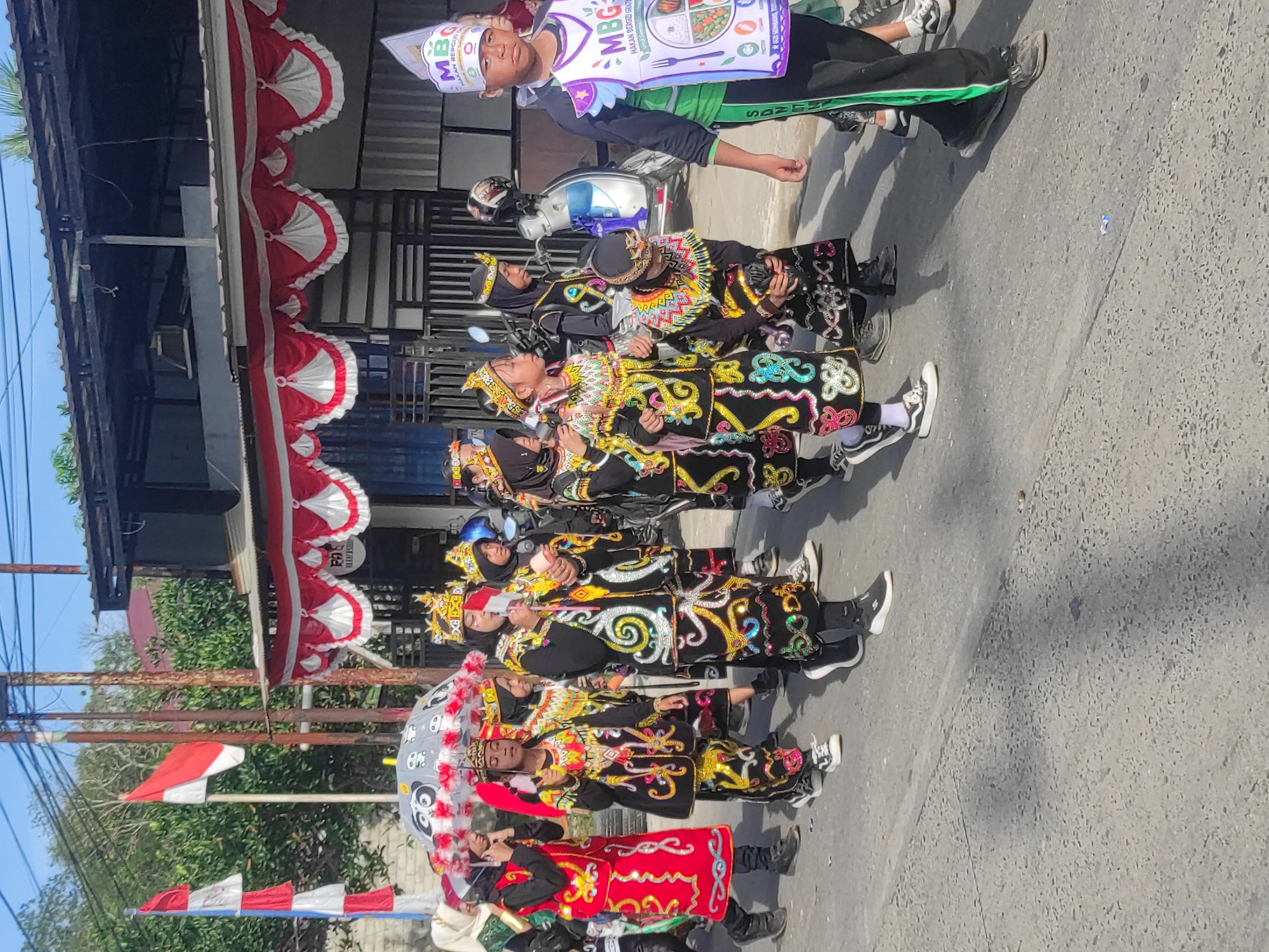A group of children in traditional attire and costumes are participating in a parade on a paved street under a clear sky. The children are adorned with ornate crowns, colorful embroidered clothing, and some are wearing headscarves. One child in the foreground is wearing a white hat with "MBG" and Indonesian text on it, and a green sash. Another child is holding a patterned umbrella. The parade is taking place in front of buildings with red and white decorative banners. Several flags, including Indonesian flags, are visible in the background. A motorcycle is parked to the right of the group. The overall scene suggests a cultural event or celebration.