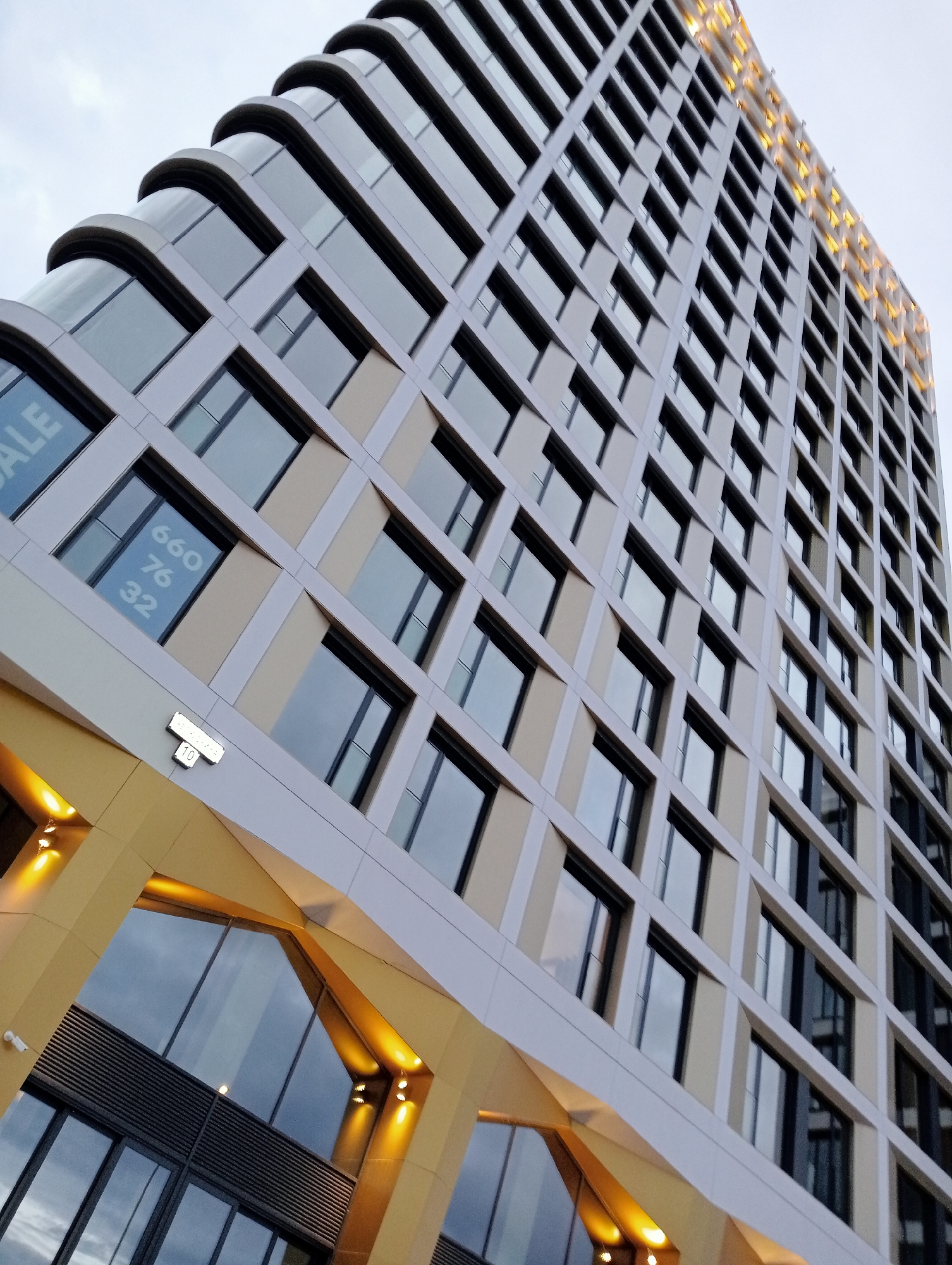 The image shows a modern skyscraper in Saint Petersburg, Russia. The building features a repeating pattern of windows and cream-colored panels. The lower portion of the building is accented with golden geometric shapes and warm lighting. In one of the windows, text reading "SALE" and numbers "660 76 32" is visible. A small plaque with the number "10" is attached to the building's facade. The sky is overcast, suggesting a cloudy day.