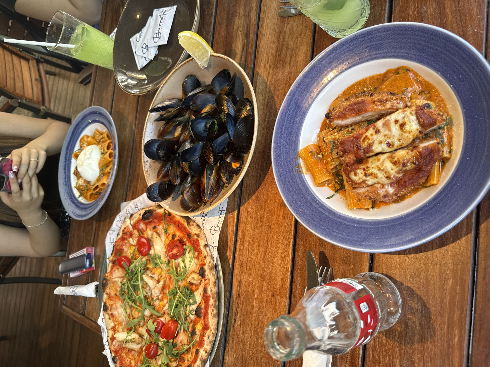 In the city of Sofia, Bulgaria, a wooden table is laden with various dishes, suggesting a meal at an outdoor restaurant. In the foreground, a clear glass bottle of Coca-Cola sits on its side, next to a fork and knife. To the right, a blue-rimmed white plate holds a pasta dish with three pieces of breaded chicken, covered in melted cheese and a red sauce, with rigatoni pasta underneath. A green beverage with a straw is visible in the upper right corner.
Further back, a pizza with arugula, cherry tomatoes, and what appears to be shredded chicken is served on a platter with a logo that reads "Burrata". Adjacent to the pizza is a bowl filled with mussels, garnished with a lemon wedge. Another white plate with pasta and a large dollop of burrata cheese is positioned to the left, with a green beverage in a tall glass with a straw nearby.
In the background, two individuals are partially visible. One person is holding a smartphone, and another is seated at the table. The overall scene depicts a casual dining experience.