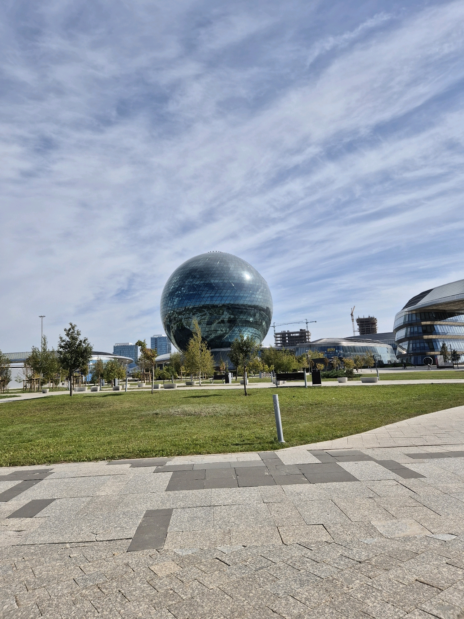 The image captures a wide-angle view of a modern city plaza with a prominent spherical glass building in the center, characteristic of Astana, Kazakhstan. The sky above is partly cloudy, with wispy white clouds streaking across a blue expanse. In the foreground, a paved walkway with a patterned stone surface leads into a well-maintained green lawn dotted with young trees and lampposts. Behind the lawn, a large, translucent, spherical building, reflecting the sky and surroundings, dominates the midground. Several other contemporary buildings with curved glass facades, and construction cranes indicating ongoing development, are visible in the background. The scene appears to be a public space, likely a park or a plaza, designed with modern architectural elements. There are no visible people in the image.