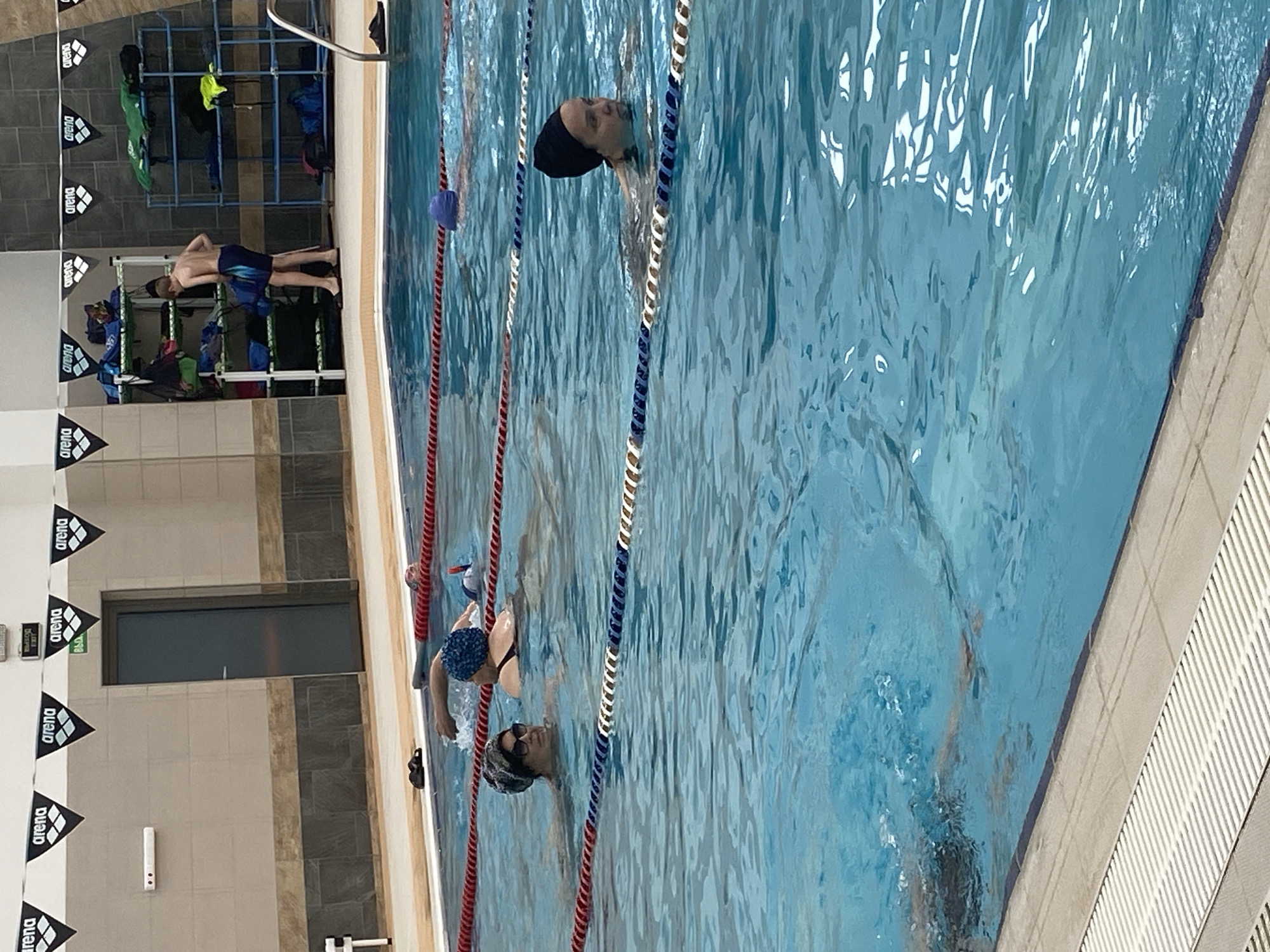 The image captures an indoor swimming pool scene. Several swimmers are in the water, separated by lane ropes marked with red, white, and blue colors. At least one swimmer is wearing a swim cap. A boy stands near a rack of equipment. The pool's edge is tiled with white and gray. Wall decorations include black and white pennants with the word "Arena." Above the pool, there is a door labeled with "Exit" in what appears to be Russian. The location is likely in Yekaterinburg, Russia.
