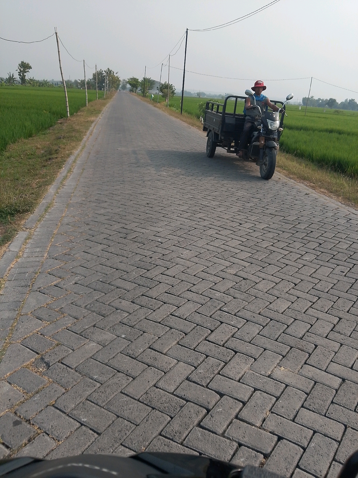 A person wearing a red hat and a blue scarf is riding a three-wheeled motorcycle down a paved road in Pangkur, Indonesia. The road is lined with green rice fields on both sides, with telephone poles and wires running along the left side of the road. The sky is hazy and light in color. The motorcycle has a cargo bed attached to the back. The road is paved with interlocking gray bricks laid in a herringbone pattern. The perspective suggests the photo was taken from another vehicle or stationary position behind the motorcycle.