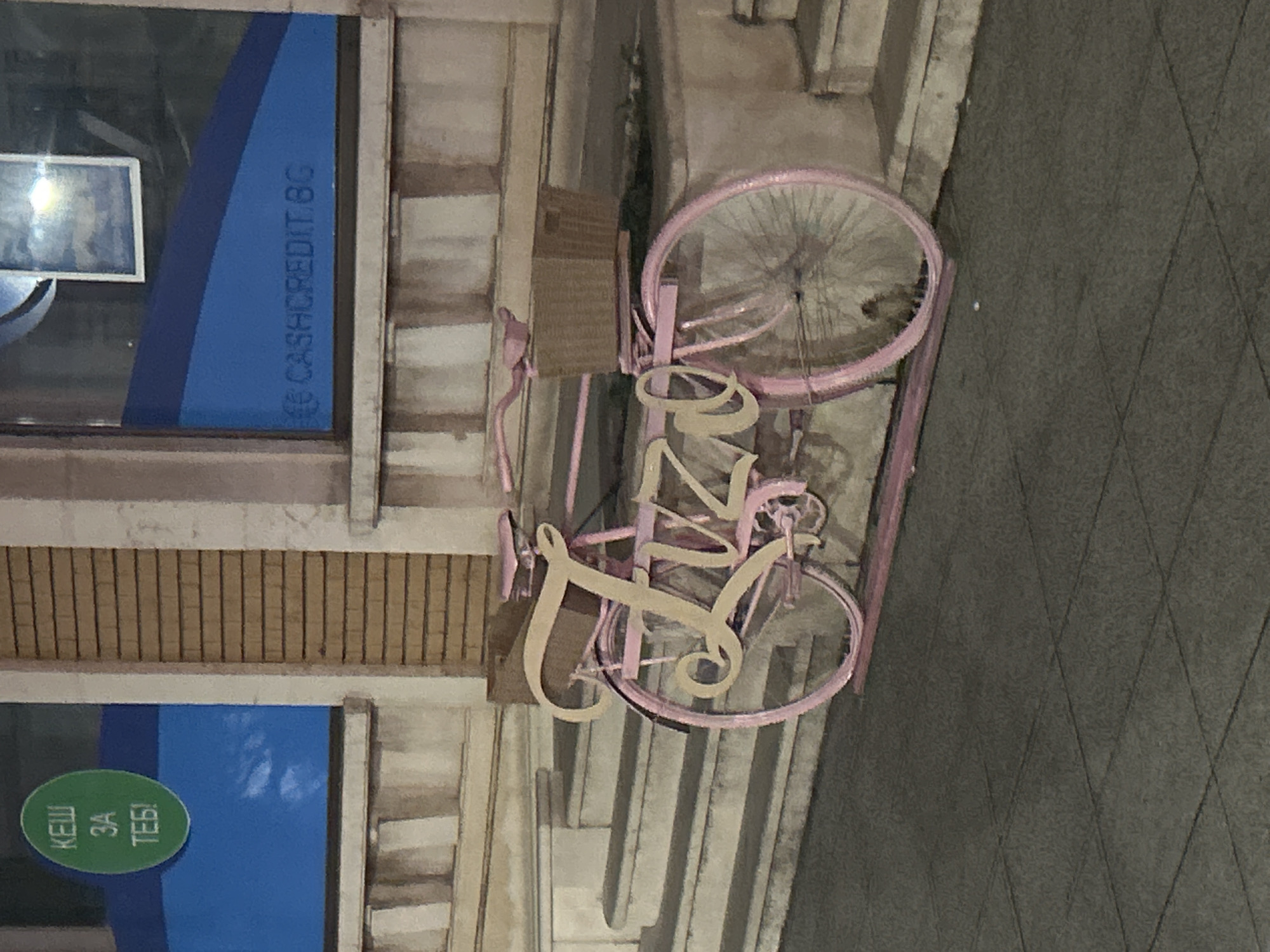 The image shows a pink bicycle with a basket, likely used as decoration for a business in Vratsa, Bulgaria. It is located outside a Cashcredit office, as indicated by the signage in the window. A sign written in Bulgarian, which translates to "Cash for you," is also visible in the window. The ground is paved.