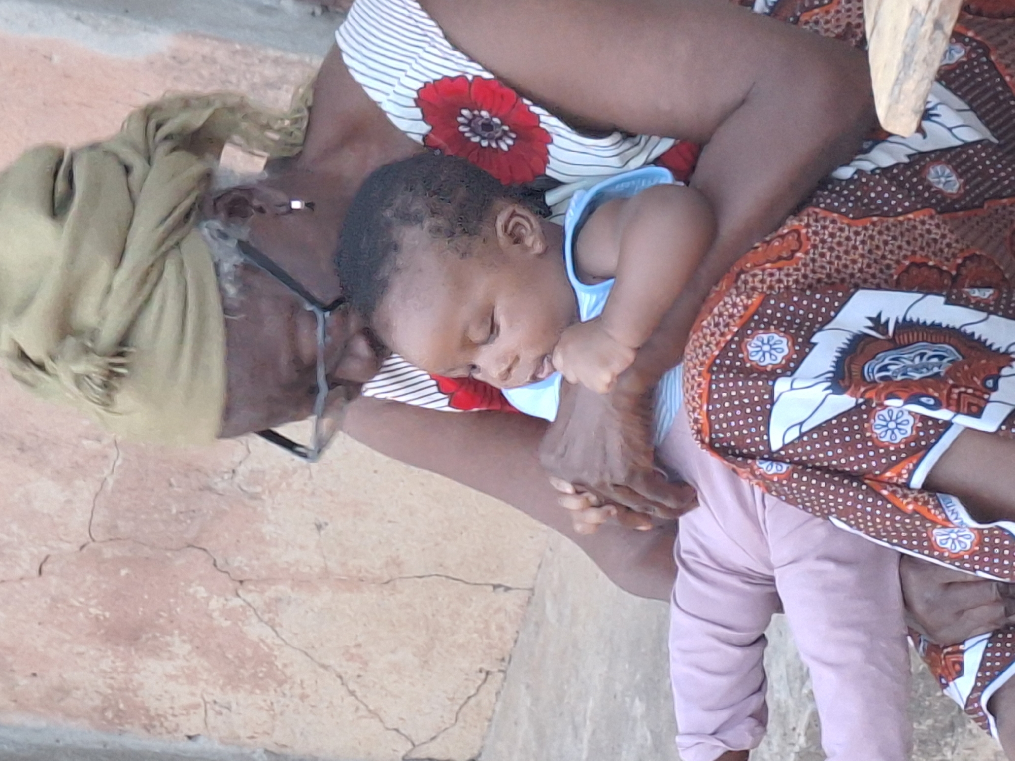 The image shows an older woman cradling a sleeping baby outdoors. The woman, with dark skin, wears a light khaki-colored headscarf and glasses. Her clothing consists of a white and black striped top adorned with a prominent red floral pattern, and a traditional patterned fabric in shades of brown, orange, and white draped over her lower body. She appears to be looking down at the baby, holding it securely.
The baby, also with dark skin, is sound asleep in the woman's arms, dressed in a light blue sleeveless top and light pink pants. One of the baby's hands is near its mouth. The setting is a cracked, light-colored concrete surface, suggesting an outdoor location, likely during the daytime. The atmosphere is peaceful and serene, highlighting a moment of comfort and tenderness between the two. The patterned fabric worn by the woman includes some small, unidentifiable white text on a blue background as part of its decorative motif. The scene is located in Sabon Tasha, Nigeria.