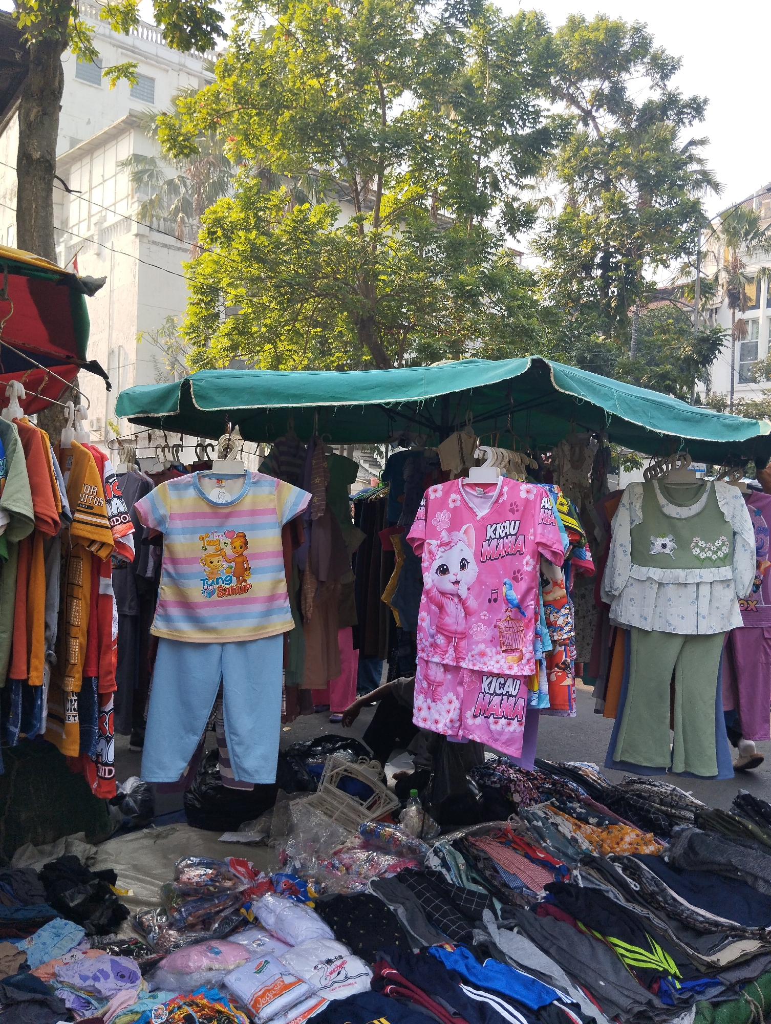 An outdoor market stall is set up on a street, likely in Surabaya, Indonesia, given the presence of children's clothing with Indonesian text. The stall is covered by a green awning, beneath which various garments are displayed. Children's clothing, including t-shirts and pants sets, are hung on hangers. In the foreground, folded clothes and packaged items are spread out on the ground. Several trees with lush green foliage are visible in the background, casting shadows on the scene. The sky is bright and appears to be daytime, with the sun illuminating the market. A white building with multiple windows is also visible behind the trees. The ground is paved, suggesting a street or sidewalk setting. Some people are partially visible in the background, with one person wearing purple pants and another person's legs visible on the right side of the frame.