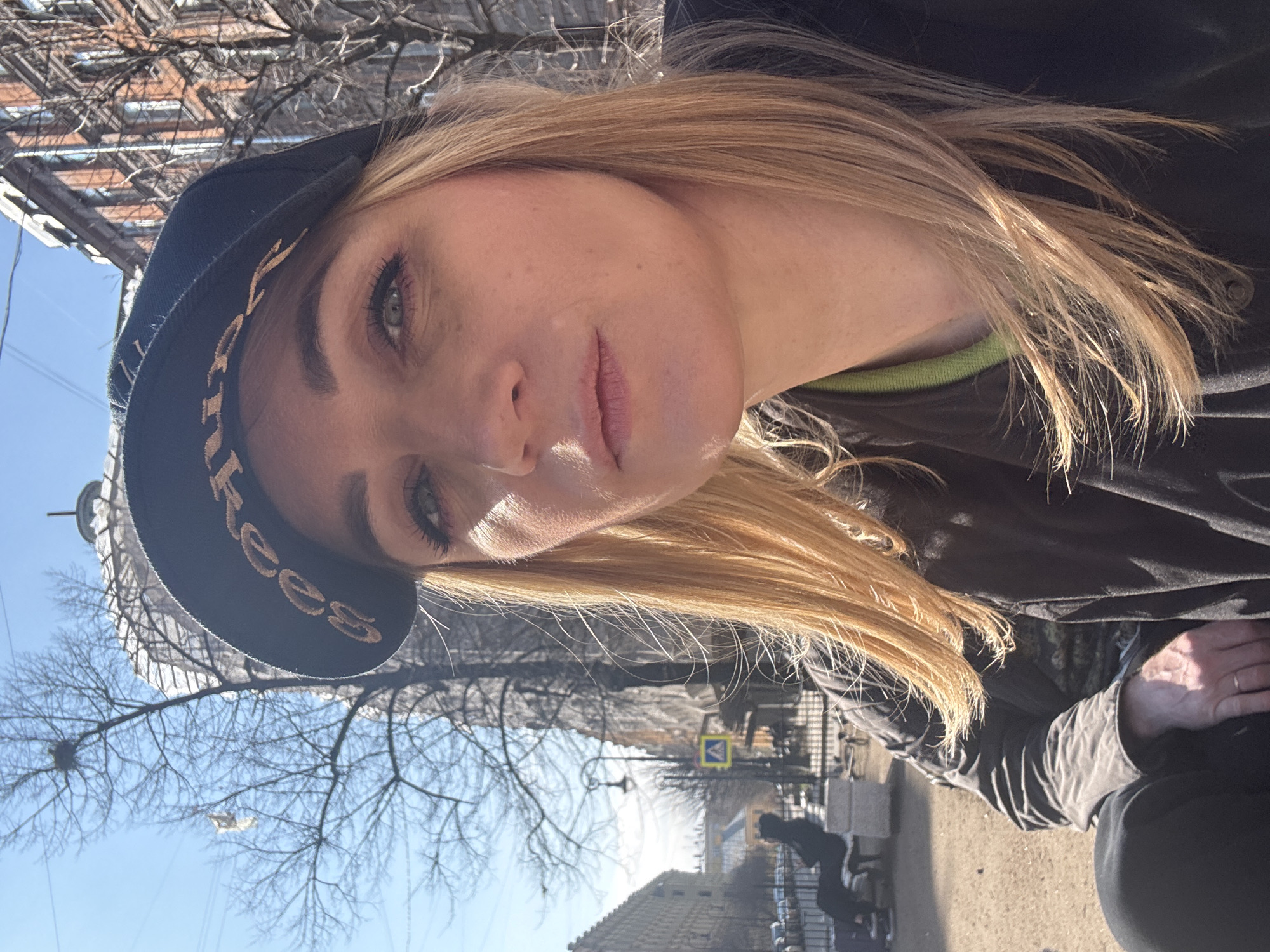 A selfie portrait features a blonde woman wearing a dark-colored hat and jacket in an outdoor setting. The hat has a logo of the letters "Kees" on the front. The woman's face is in sharp focus, revealing details such as her skin tone and eye color. The background is blurred, but it includes elements of a city street, trees, and buildings. A road sign is visible. The buildings appear to be of historical architecture, suggesting that the location may be within the city of Saint Petersburg, Russia. People are visible in the background, some seated.