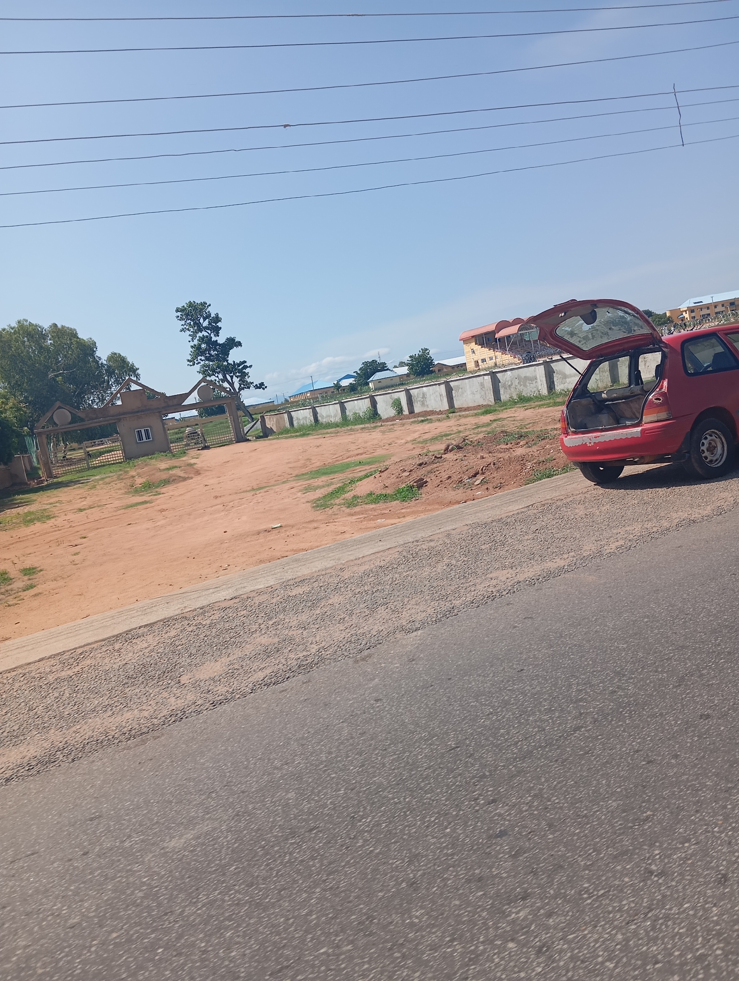 The image depicts an outdoor scene in Jalingo, Nigeria. The foreground is dominated by a paved road, with a reddish-brown dirt area to the left, separated by a curb. Beyond the dirt area, there's a wrought-iron gate with an ornate archway, suggesting an entrance to a property. A small, single-story building with a white-framed window is visible within the gated area, along with some greenery and trees.
To the right of the frame, a red station wagon with its hatchback open is parked on the dirt area. The car appears to be older, and its interior is partially visible. In the background, a series of concrete walls line the property, and beyond that, several multi-story buildings are visible, some with light-colored facades and windows. A few scattered trees and vegetation are also present. The sky is clear and blue, indicating a sunny day. Multiple power lines stretch across the sky. The scene appears to be set during daylight hours.