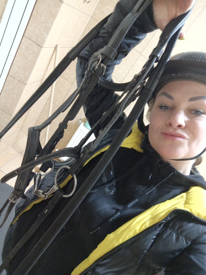 A female subject is depicted in an indoor setting, oriented diagonally across the frame. She has light-colored hair, partially visible beneath a dark, textured riding helmet. Her facial expression shows pursed lips as she looks directly towards the camera. She wears a black puffer jacket featuring distinct yellow panels on the shoulders and upper sleeves, over a dark undergarment. Her left hand, with yellow-painted fingernails, is raised and holds a dark leather horse bridle equipped with metallic rings and a snaffle bit. The background consists of light brown/beige rectangular tiles, arranged diagonally, with a white wall-mounted radiator visible in the lower-left. A light-colored wooden plank or beam is also present diagonally in the upper-left background. The specific city location of Gorka, Russia, is not discernible from the visual information provided in the image.