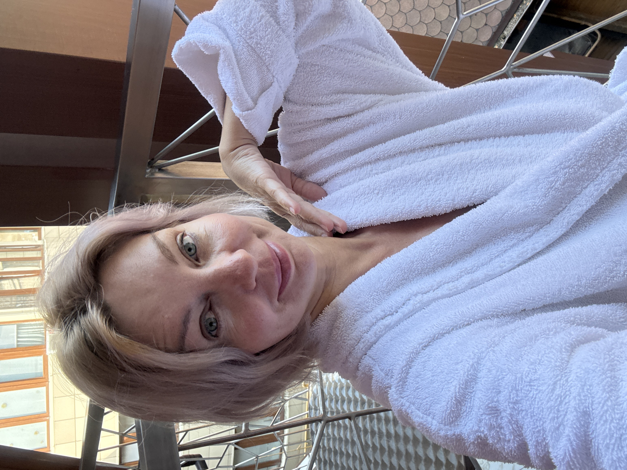 A woman is posing for a selfie outdoors in Estosadok, Russia. She is wearing a white bathrobe and has short, light-colored hair with pinkish undertones. Her left hand is resting on her neck and shoulder, and she is smiling faintly at the camera. In the background, there is a metallic railing, a building with wooden window frames, and a textured wall.
