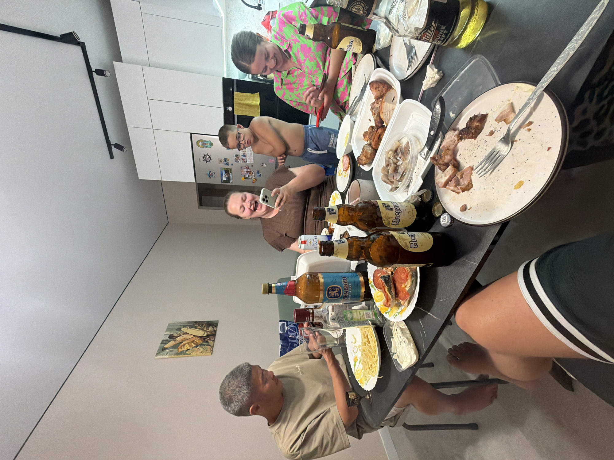 The image depicts a group of four people, two adults and two children, gathered around a table laden with food and drinks, likely in a residential setting. The environment appears to be an indoor dining area, possibly a kitchen or living room, with light-colored walls and modern cabinetry visible in the background. A track lighting system is installed on the ceiling.
On the table, there are several plates of food, including what appears to be pasta, fish, and roasted meats. Various bottles of alcoholic beverages, including beer and liquor, are present, alongside glasses and cutlery. The people are engaged in conversation and interacting with their mobile phones. The overall atmosphere suggests a casual gathering or meal.
The image does not contain any explicit visual cues to identify the specific location within the city of Novorossiysk, Russia.