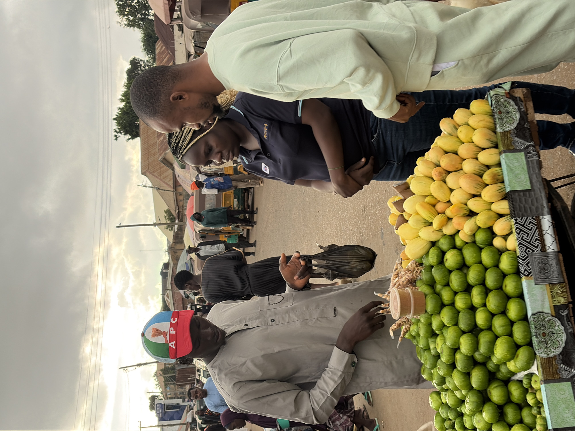 In Gura Top, Nigeria, three people are gathered around a fruit stall. A man in a grey traditional outfit and a red and white cap with an "APC" logo is holding a black plastic bag and interacting with the fruit. To his right, a woman with braided hair, wearing a dark polo shirt and jeans, stands with her hands clasped. Beside her, a man in a light green sweatshirt and matching pants looks towards the fruit stall. The stall is laden with a large pile of green fruits, appearing to be some type of citrus, and a smaller pile of yellow fruits, possibly mangoes. Two small plastic containers with a brown substance are placed on top of the green fruit. The background shows a busy market scene with other people, vehicles resembling auto-rickshaws, buildings with corrugated metal roofs, and overhead power lines under a cloudy sky.