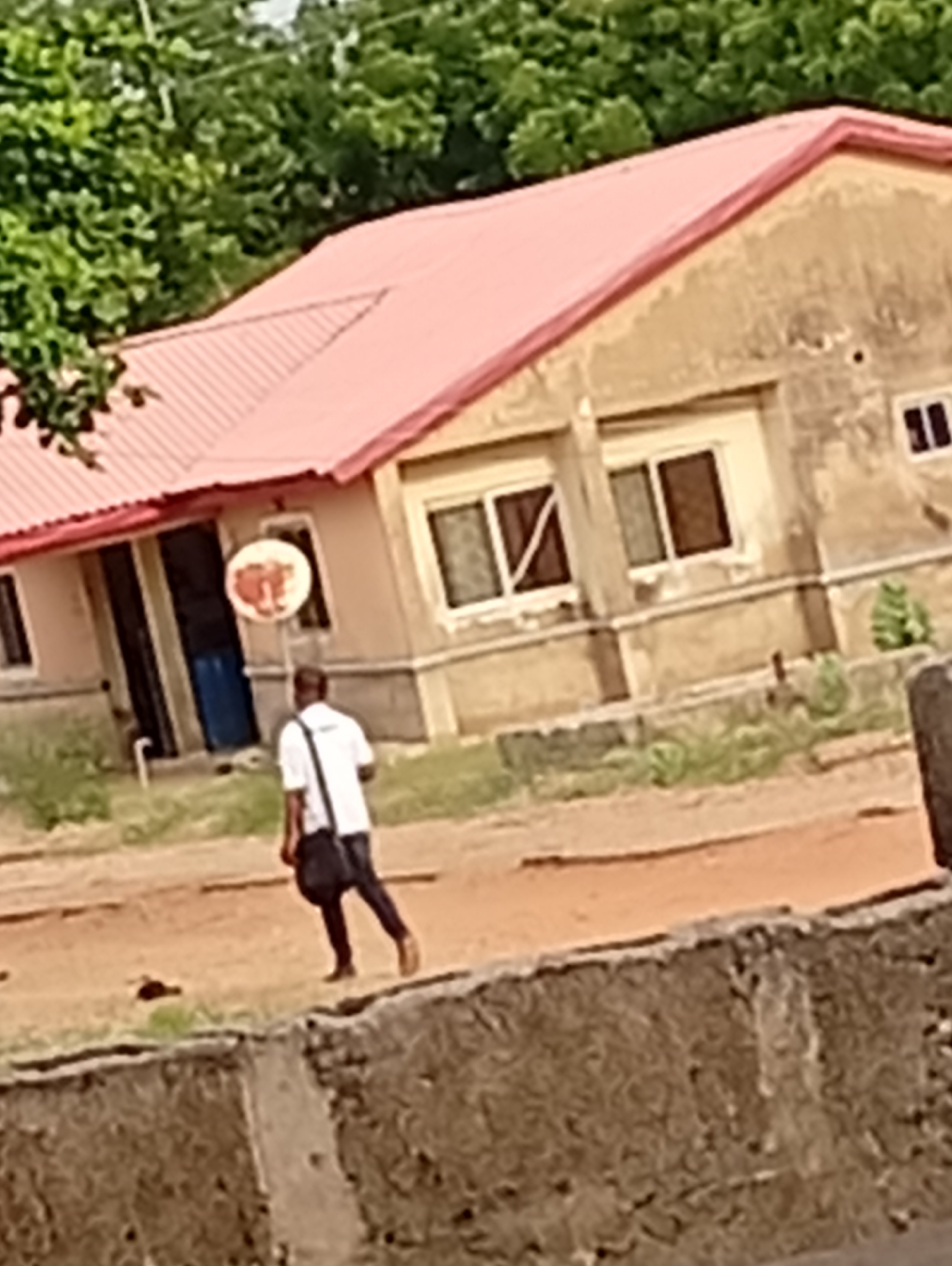 A person is walking on a dirt road in front of a beige, one-story building with a red roof in Unknown, Unknown. The building has multiple windows and what appears to be a satellite dish on the exterior wall. The road is bordered by a low, concrete wall in the foreground. Trees are visible in the background. The person is wearing a light-colored shirt, dark pants, and carrying a dark bag.